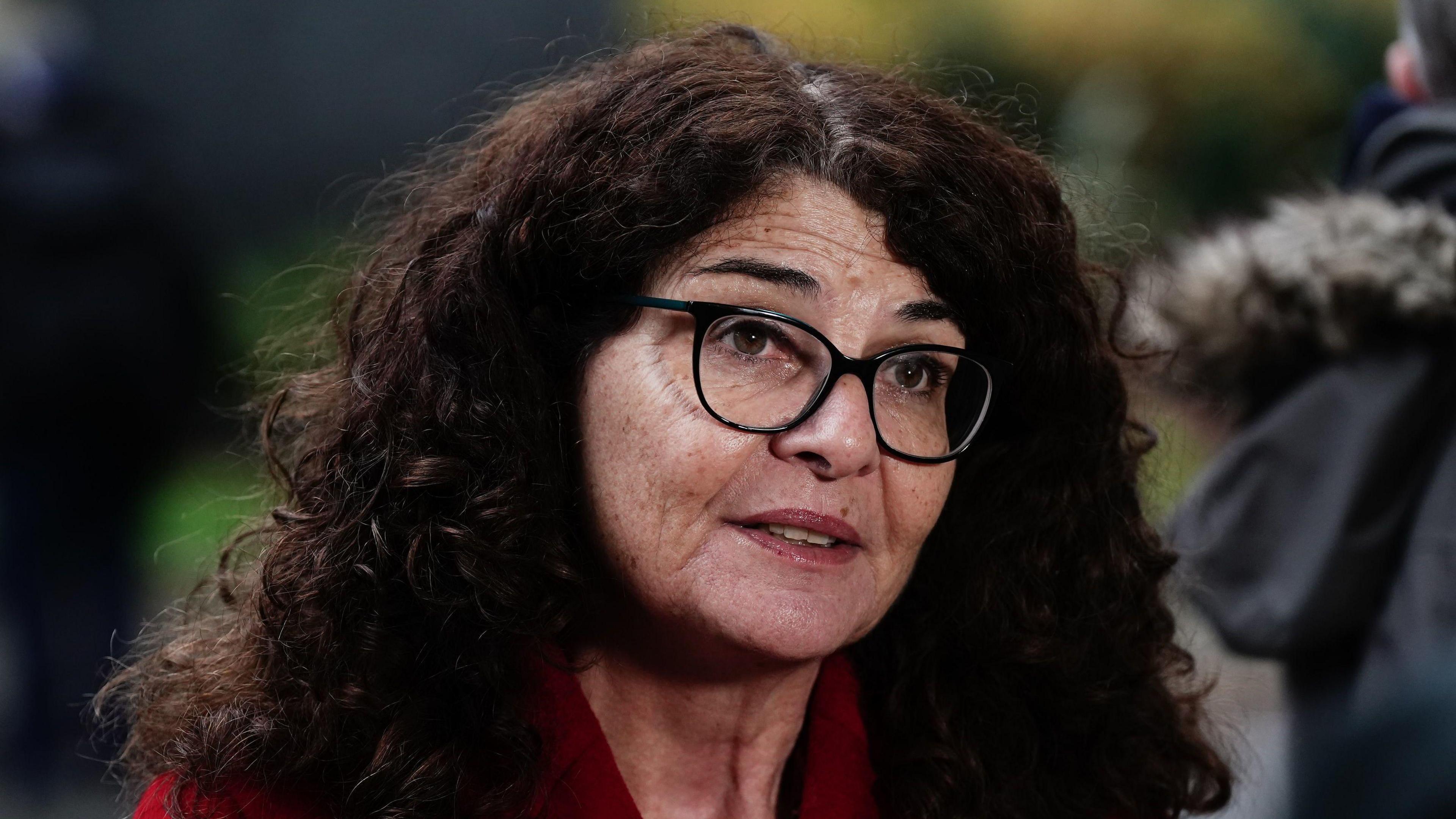 A woman with long curly hair wears glasses and a red top. Blurred in the background are some trees. 