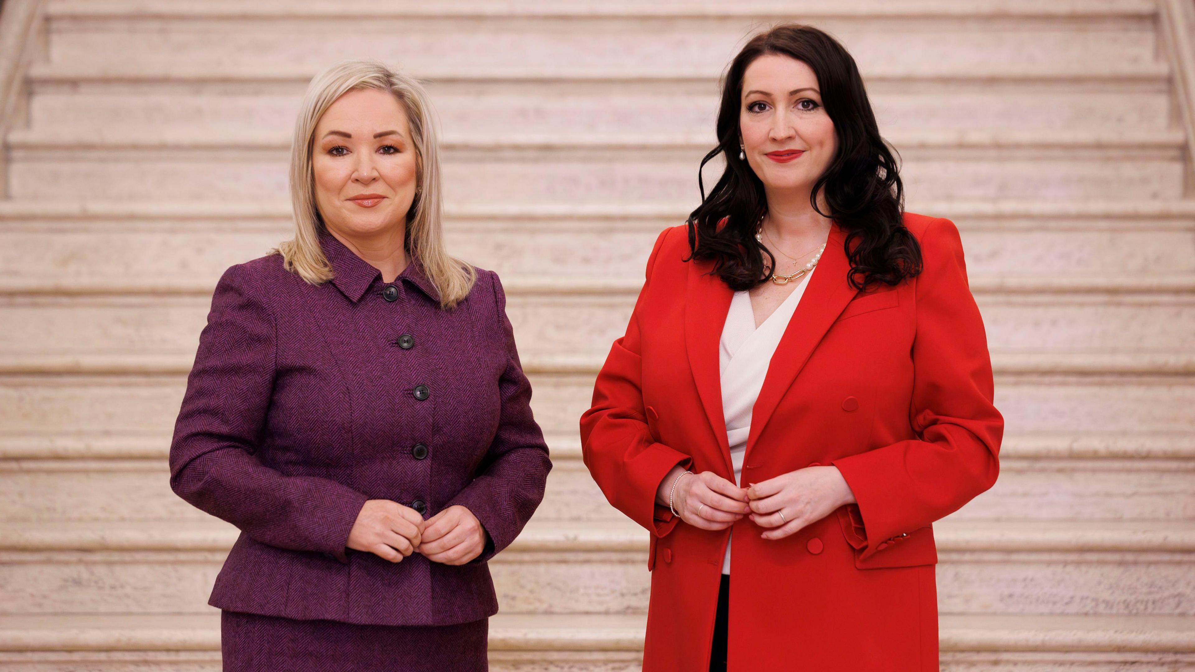 Two women are in the frame smiling to camera. On the left is a woman with blonde, shoulder length hair wearing a purple skirt suit. On the right is a dark haired woman wearing a red blazer, white shirt and black trousers.