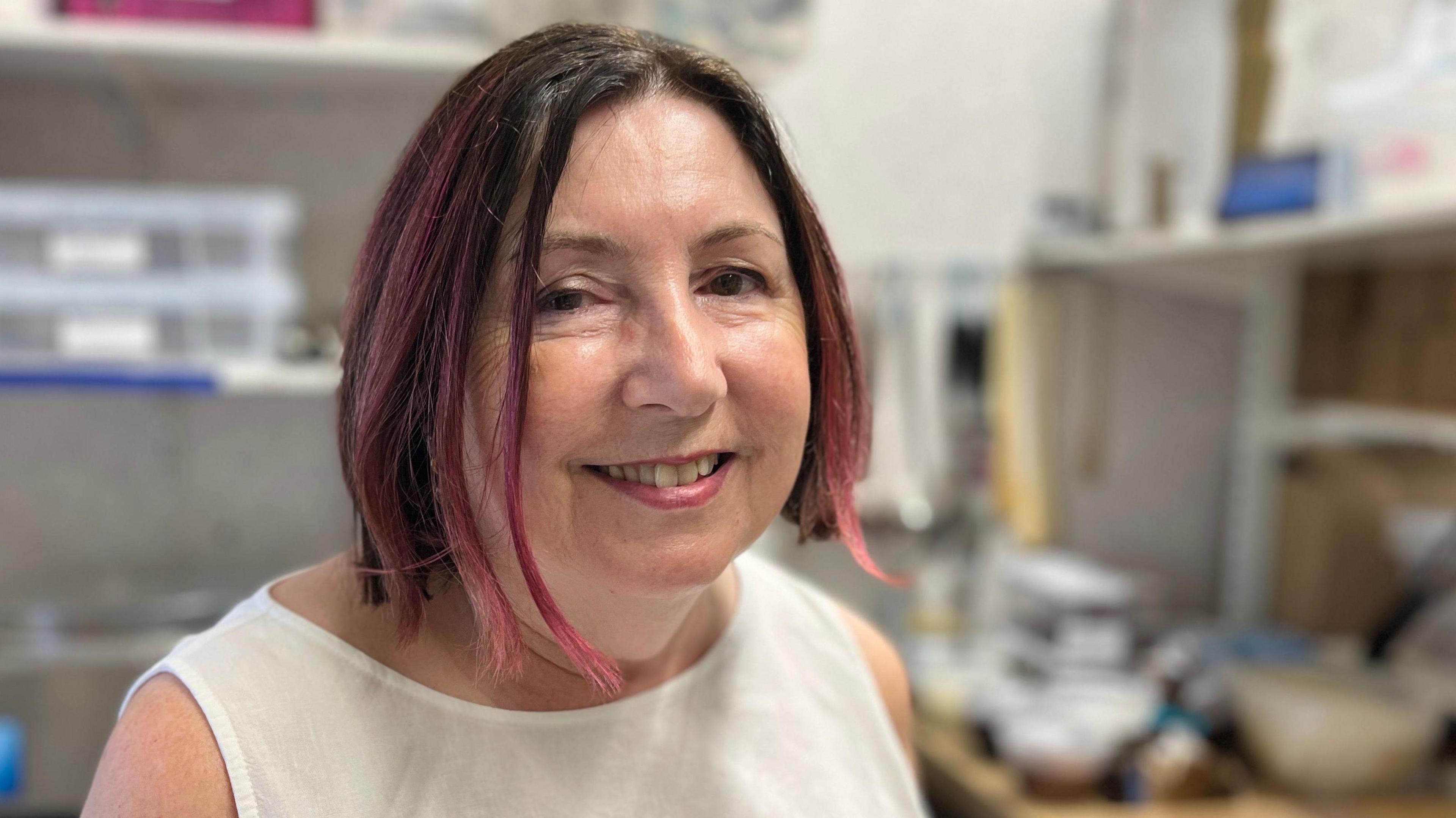 Isabelle Chappelle has short brown and pink hair and is wearing a cream short sleeved T-shirt. She is sitting in a room which has several plastic containers on shelves.