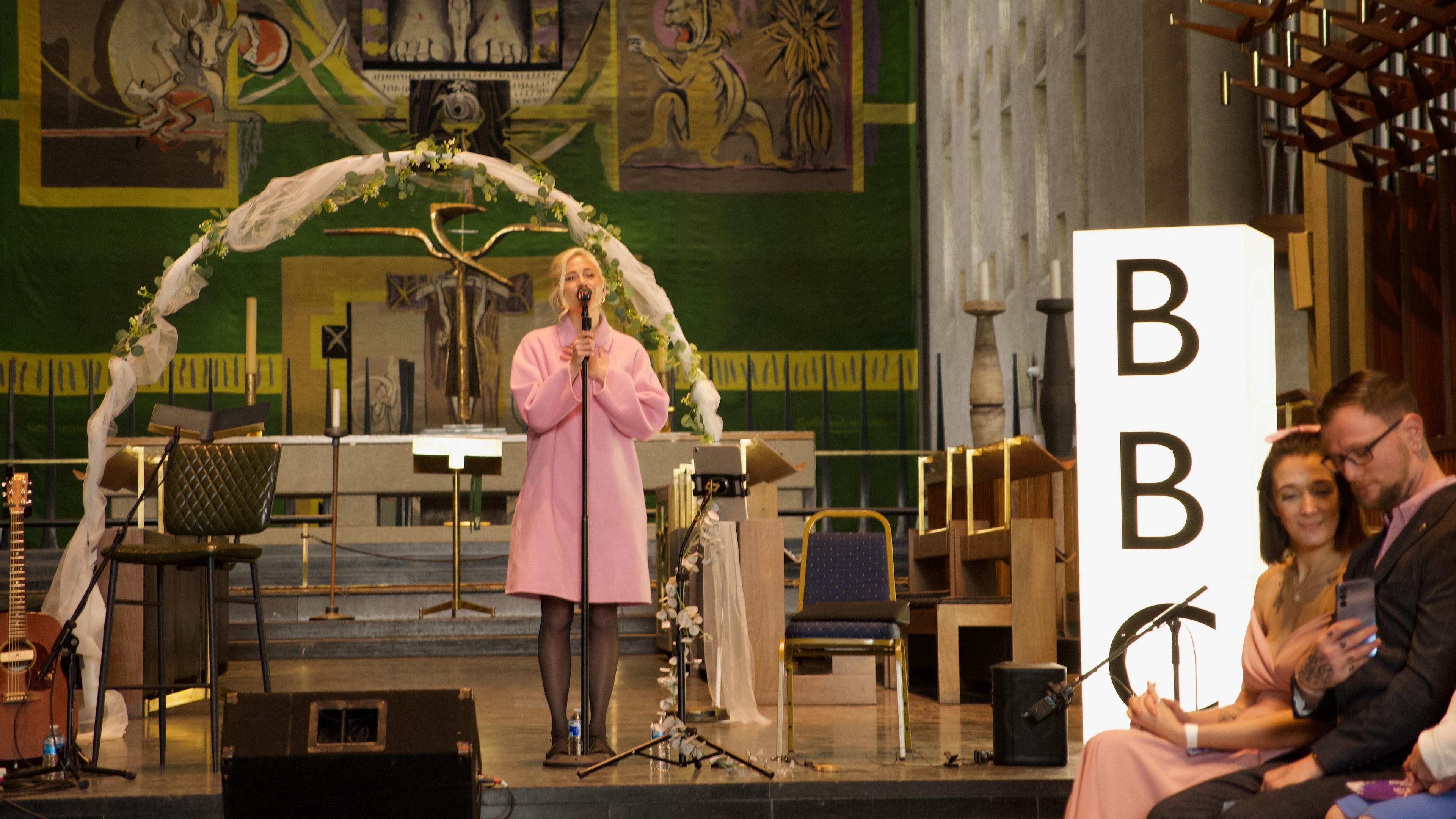 Singer in a pink coat performs at Coventry Cathedral. On the right a couple are taking a selfie to incorporate them and the singer