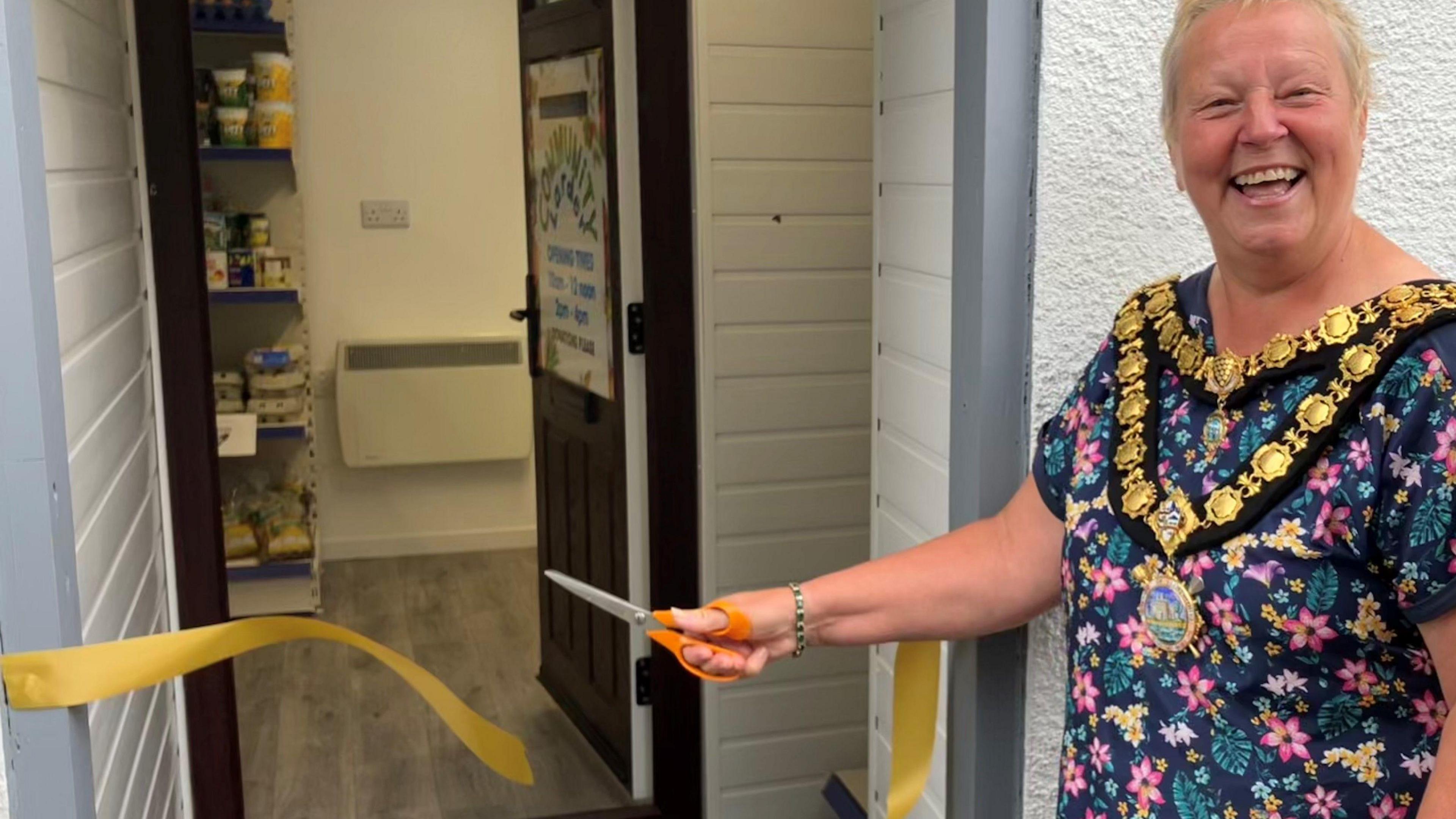 Mayor of Lostwithiel Karin Henderson, wearing a floral dress and her ceremonial chains, cuts a yellow ribbon across a door to pronounce the opening of the community larder