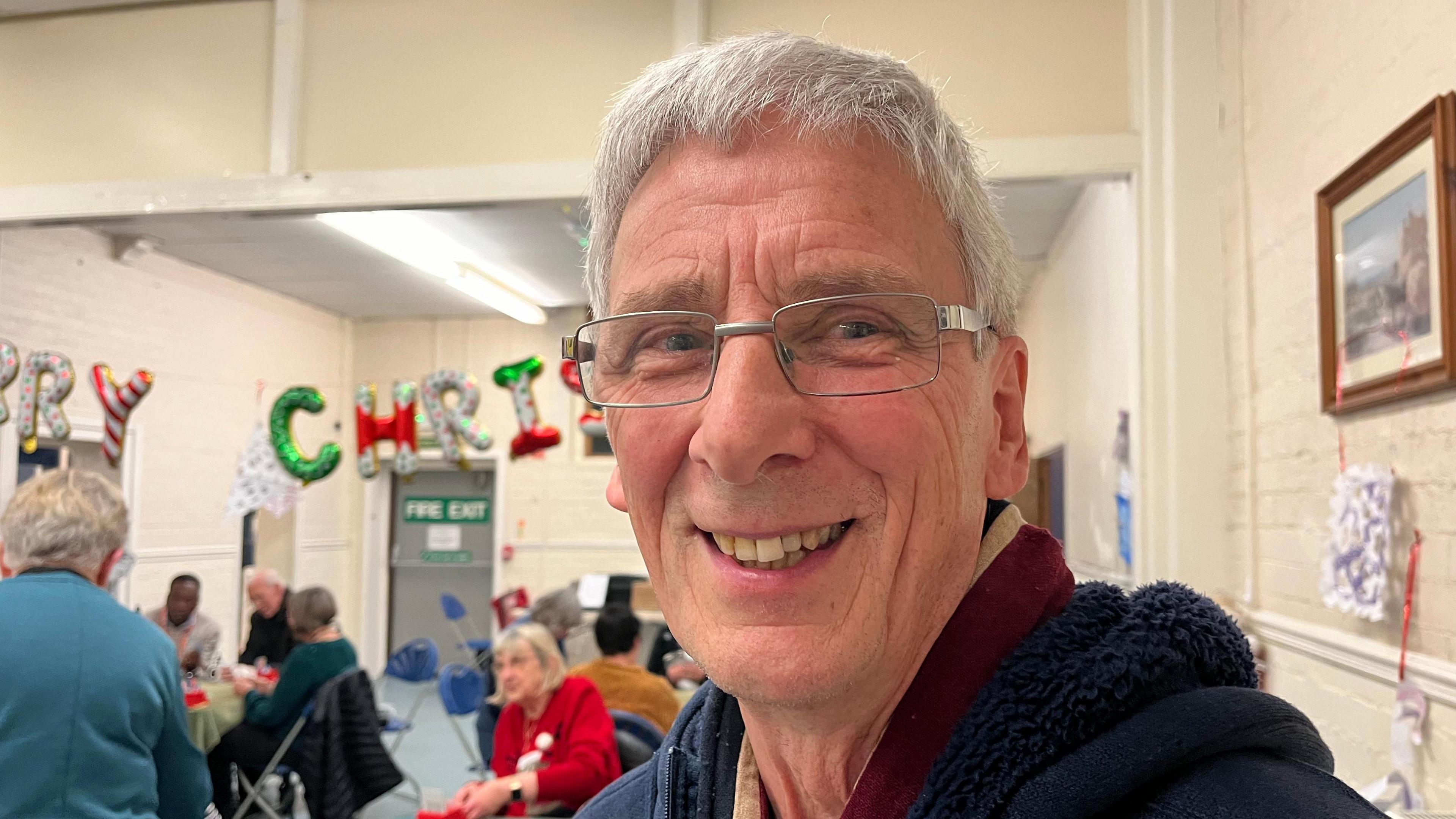 Malcolm looking into the camera, over his right shoulder you can see tables being set up for bridge. He's stood in a village hall-type building, and you can see a banner that reads 'Merry Christmas' spelled out by brightly coloured balloons.