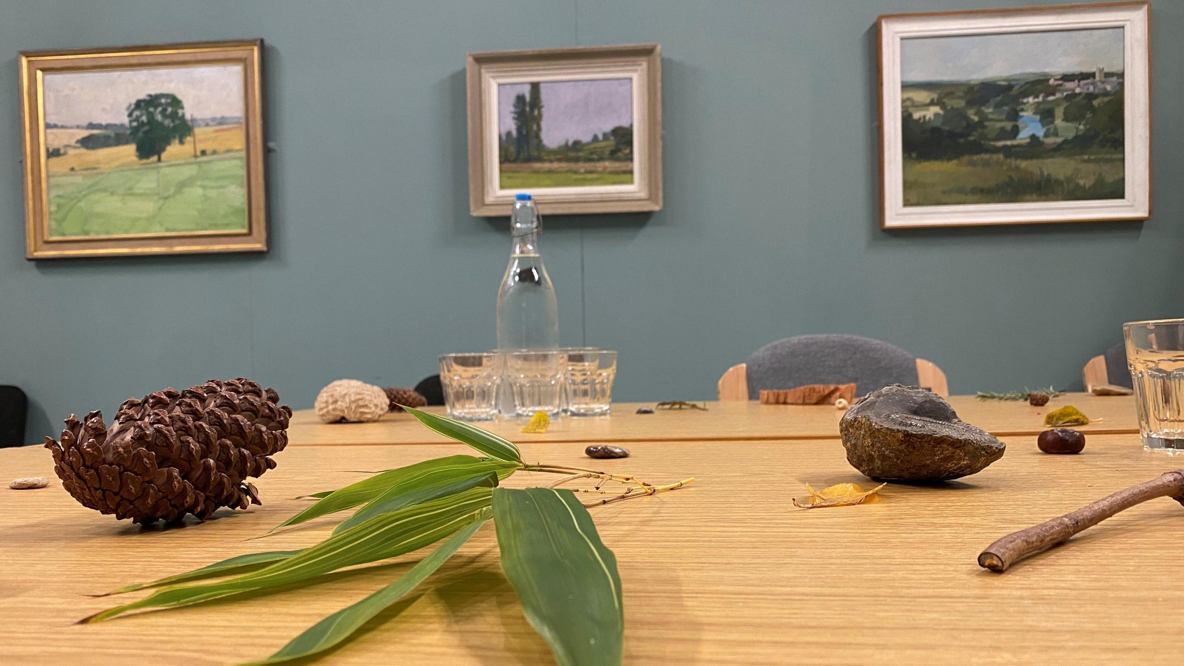 A table in a meeting room covered with items from the natural world such as acorns, twigs and rocks. There are also glasses and a water bottle on it. Some pictures are hanging on the green wall behind it.