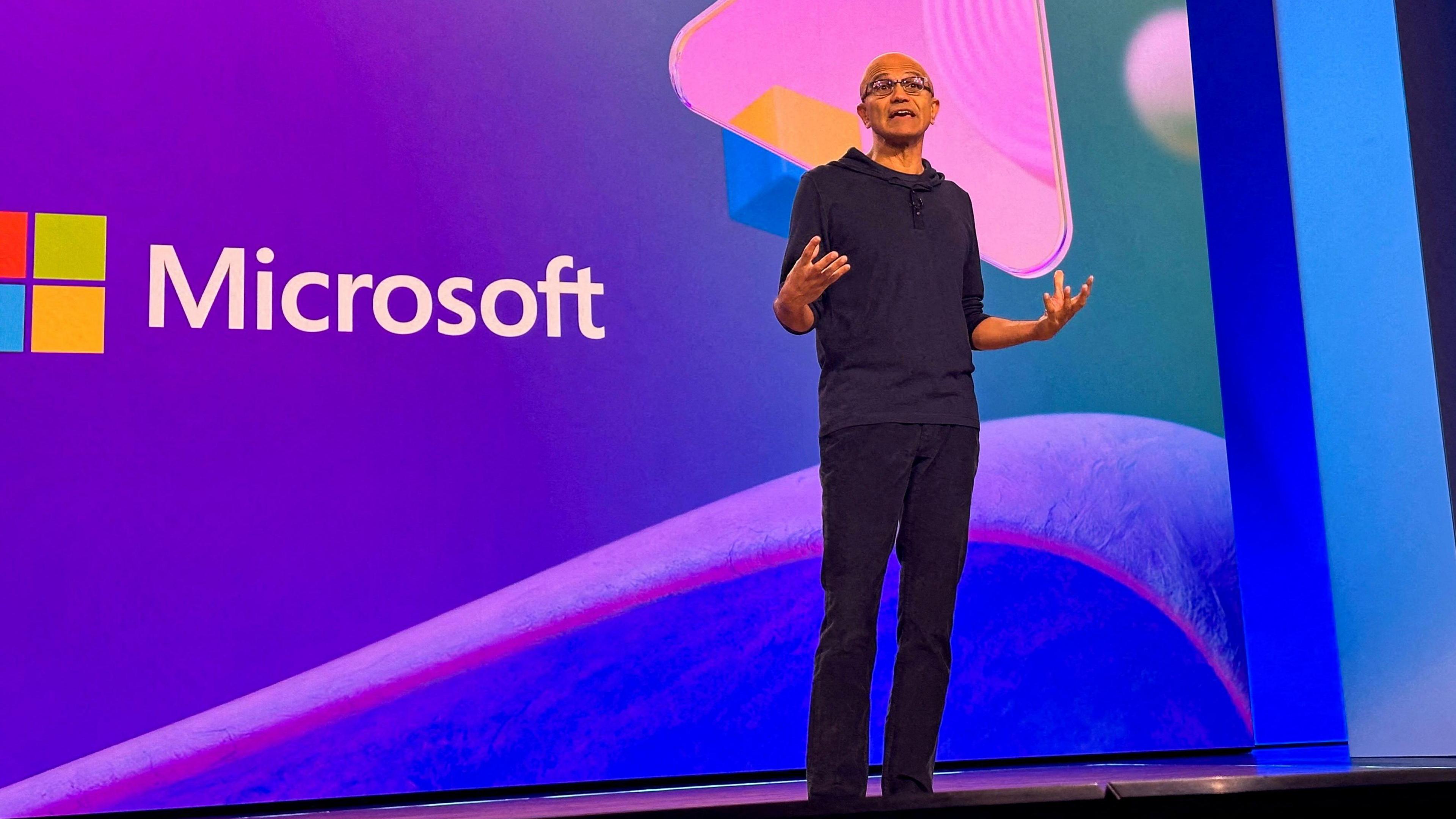 A man wearing black clothing speaks in front of a crowd, with a logo reading "Microsoft" in the background.