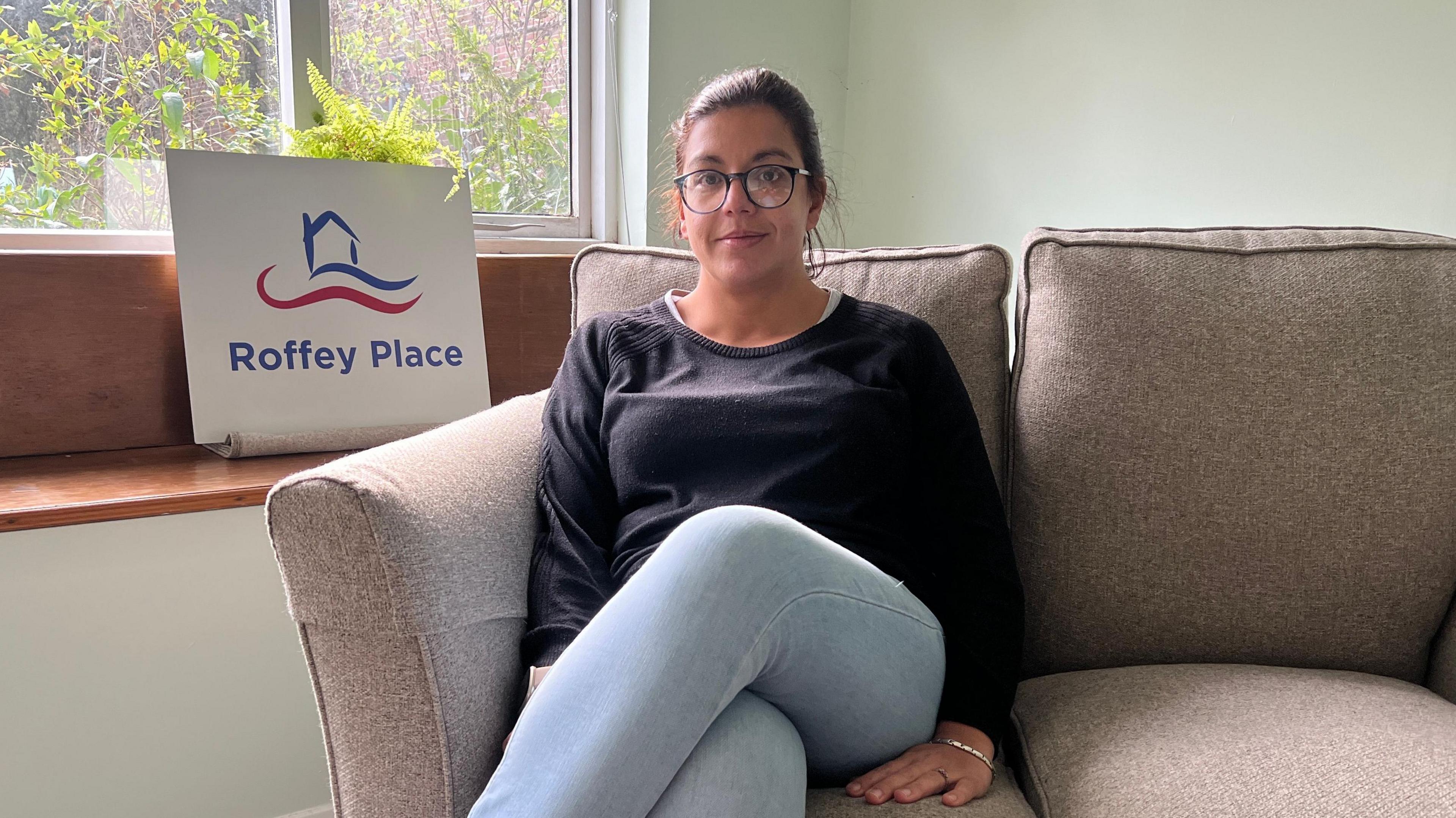 Tara Sims sitting on a break sofa. She is wearing glasses, blue jeans and a black jumper. She is looking directly at the camera, with a sign reading Roffey Place on it behind her. 