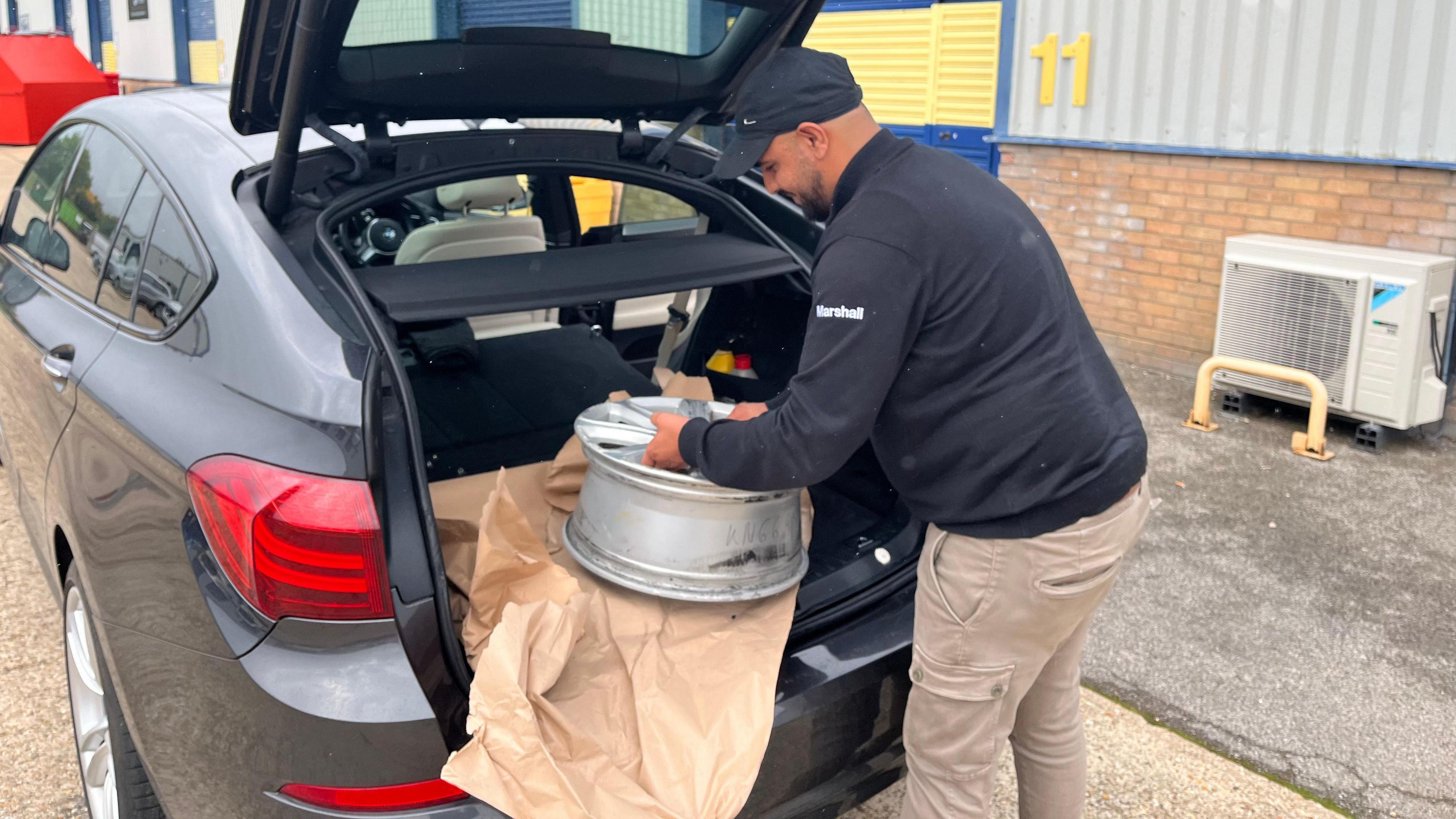 A customer reaches into his boot to lift a damaged alloy wheel. He is wearing a dark cap and jumper and beige trousers. The car is grey and there is brown paper in the boot between the alloy and the car's interior. 