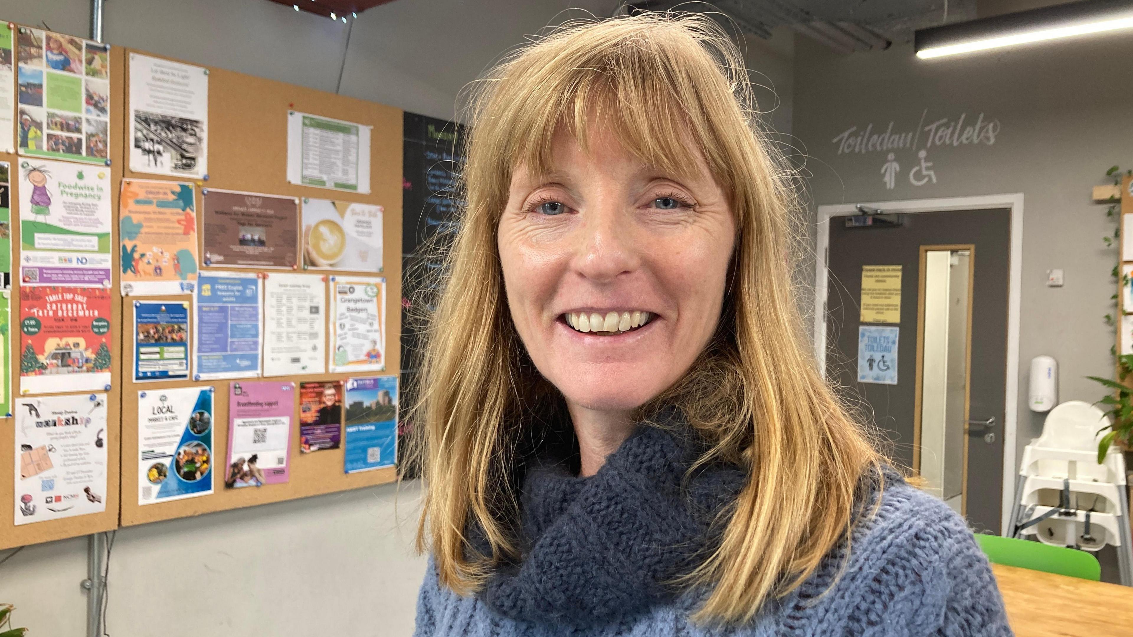 Dr Sarah Fry at a community centre in Grangetown. She is smiling and has shoulder length blonde hair. She is wearing a blue wool polo neck jumper and over her right shoulder is a noticeboard featuring numerous flyers.