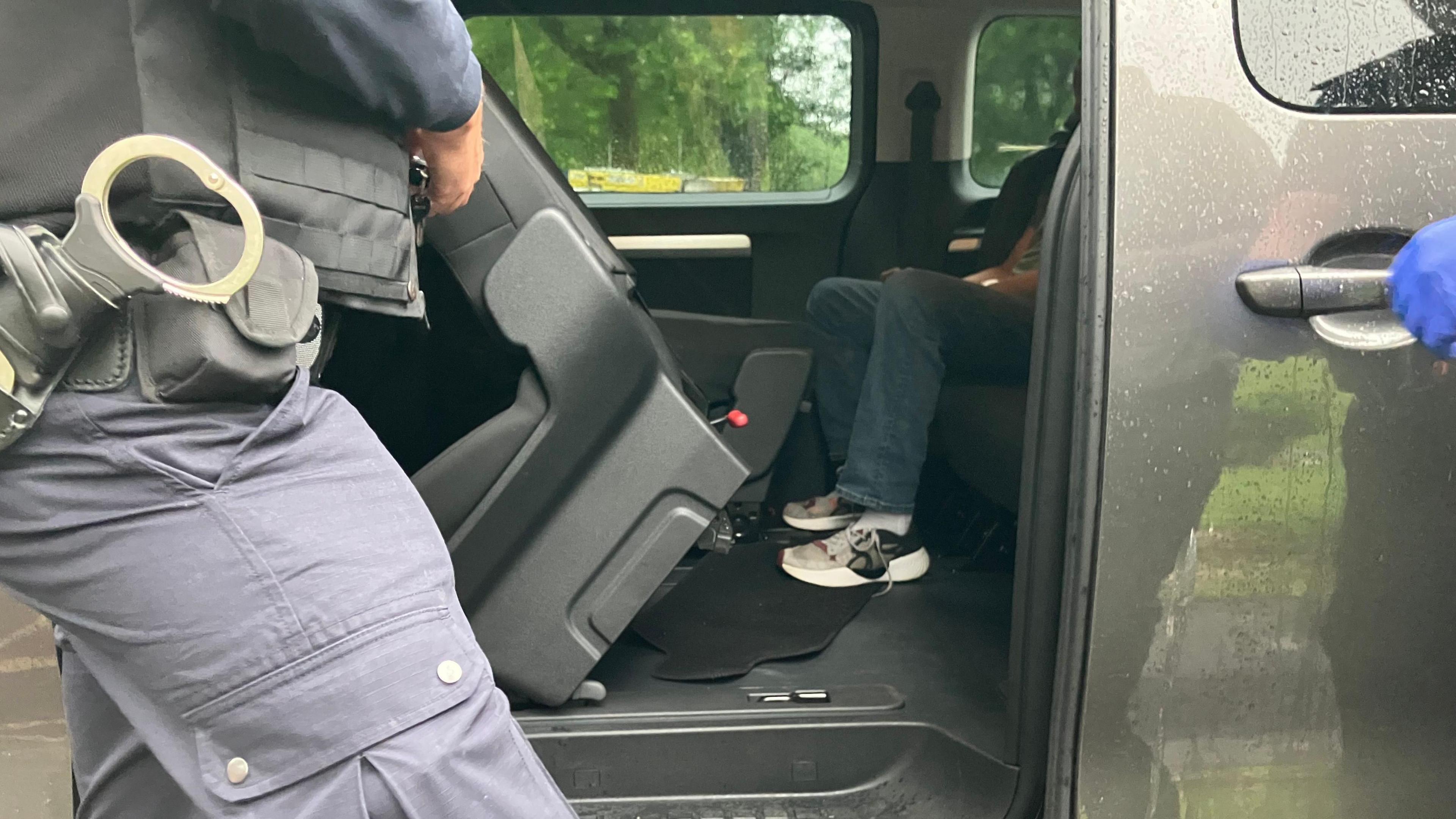 A man sits in the back of a grey van with his hands resting on his thighs. A person stands in front of the open door, with handcuffs attached to a belt around their waist. Another person has their gloved hand on the handle of the van door.