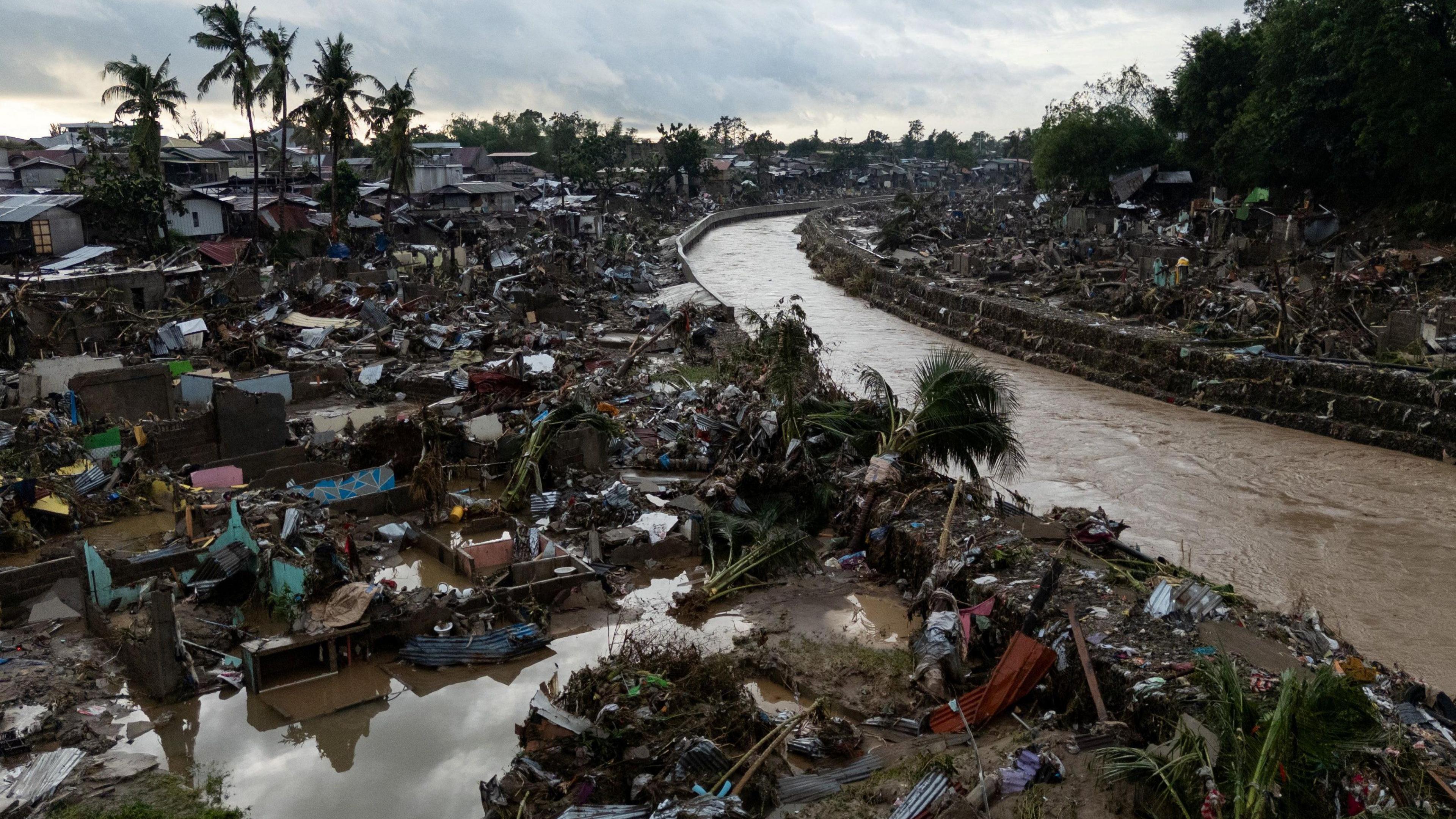 Drone view of the damage caused by Typhoon Kalmaegi in Talisay, Cebu, Philippines. There is a brown river flowing. Either side of the river you can just see debris covering the ground.