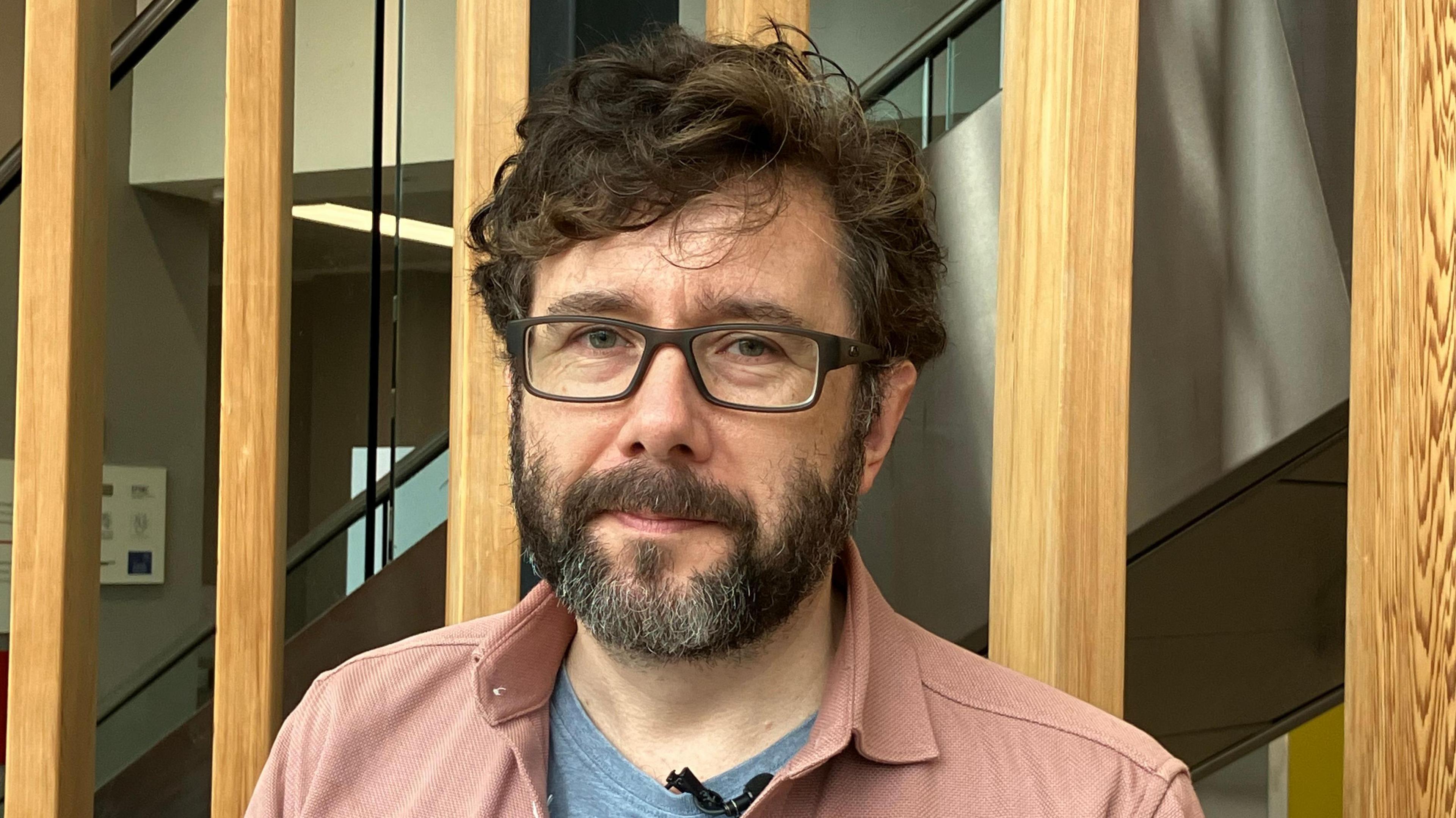 Mae Dr Rhys Bevan Jones yn edrych yn syth i'r camera. Mae ganddo wallt tywyll cyrliog a barf. Mae ganddo sbectol ac mae'n gwisgo crys pinc.