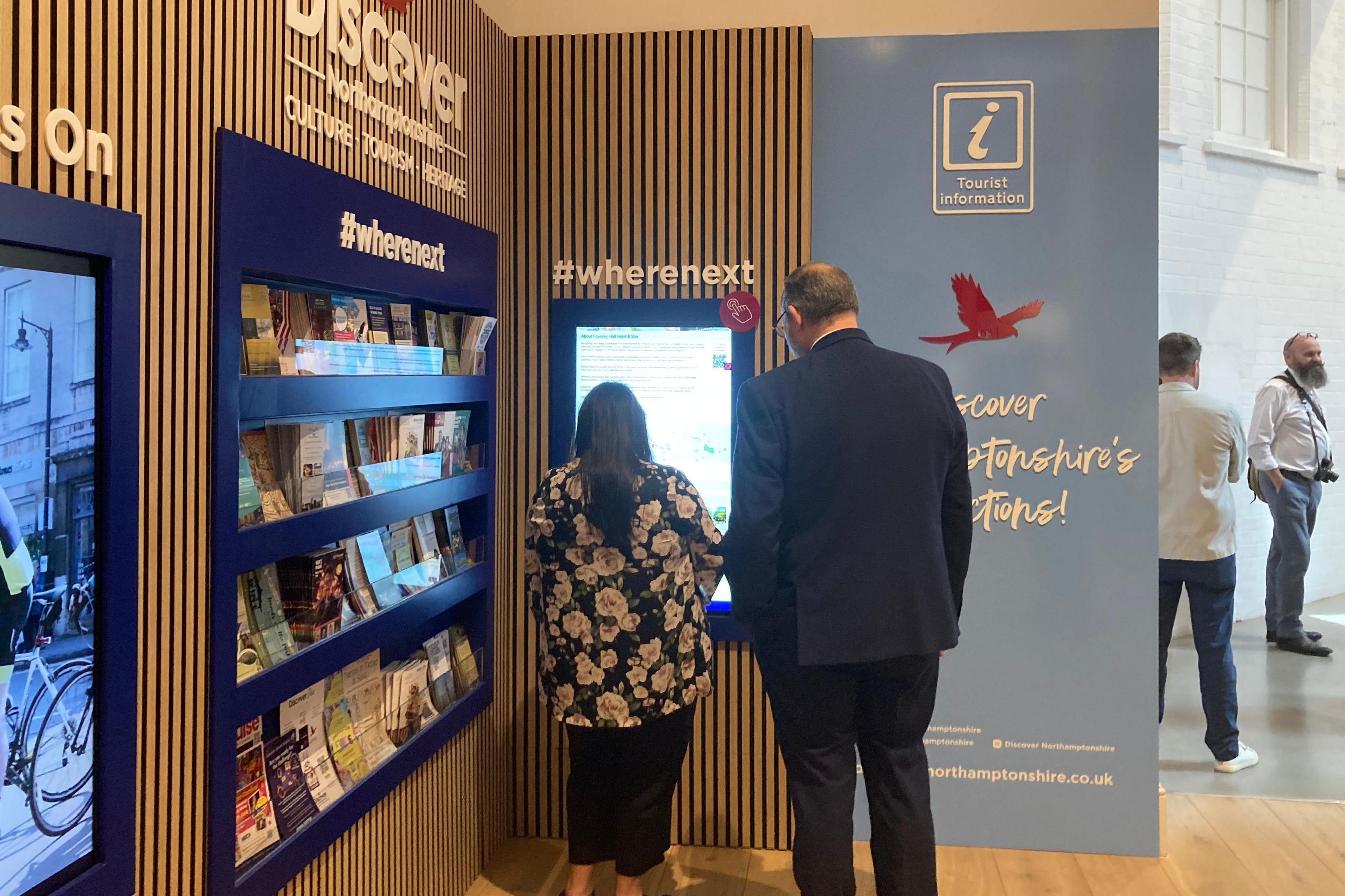 The back of a tall man in a dark suit and a person with long hair and a floral top, as they look at the new digital screen at the information point indoors.