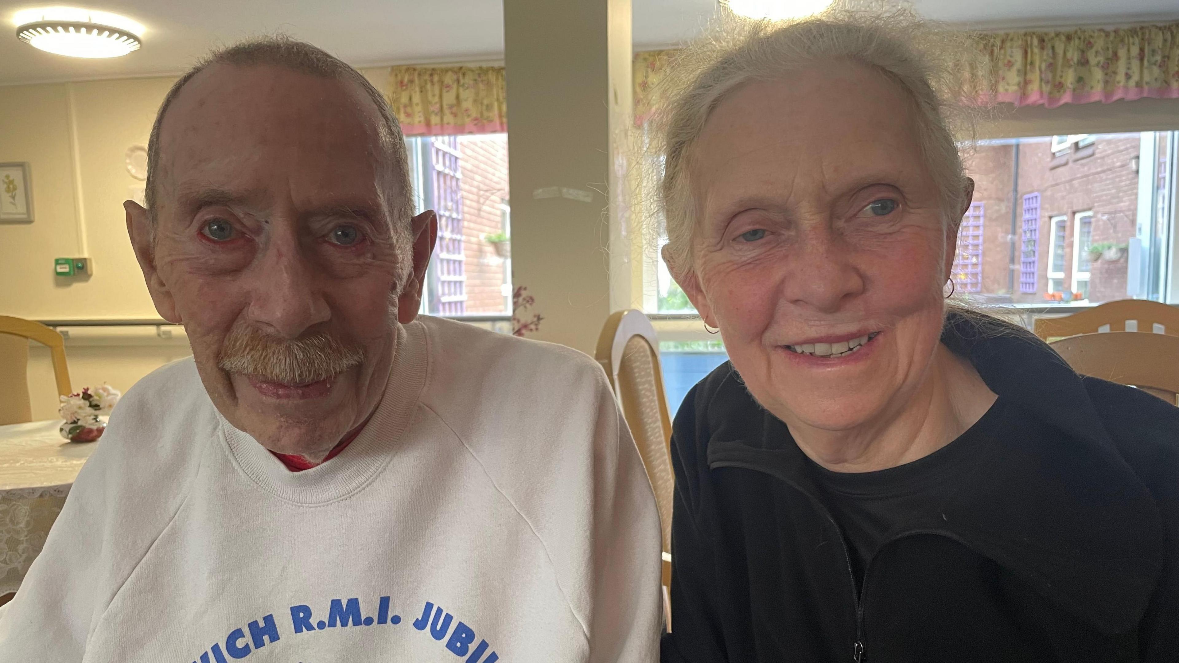 An image of John Payn and his daughter Christine Sweatman, he is wearing a white jumper with blue writing on it, she is wearing a black fleece.  Both are smiling.
