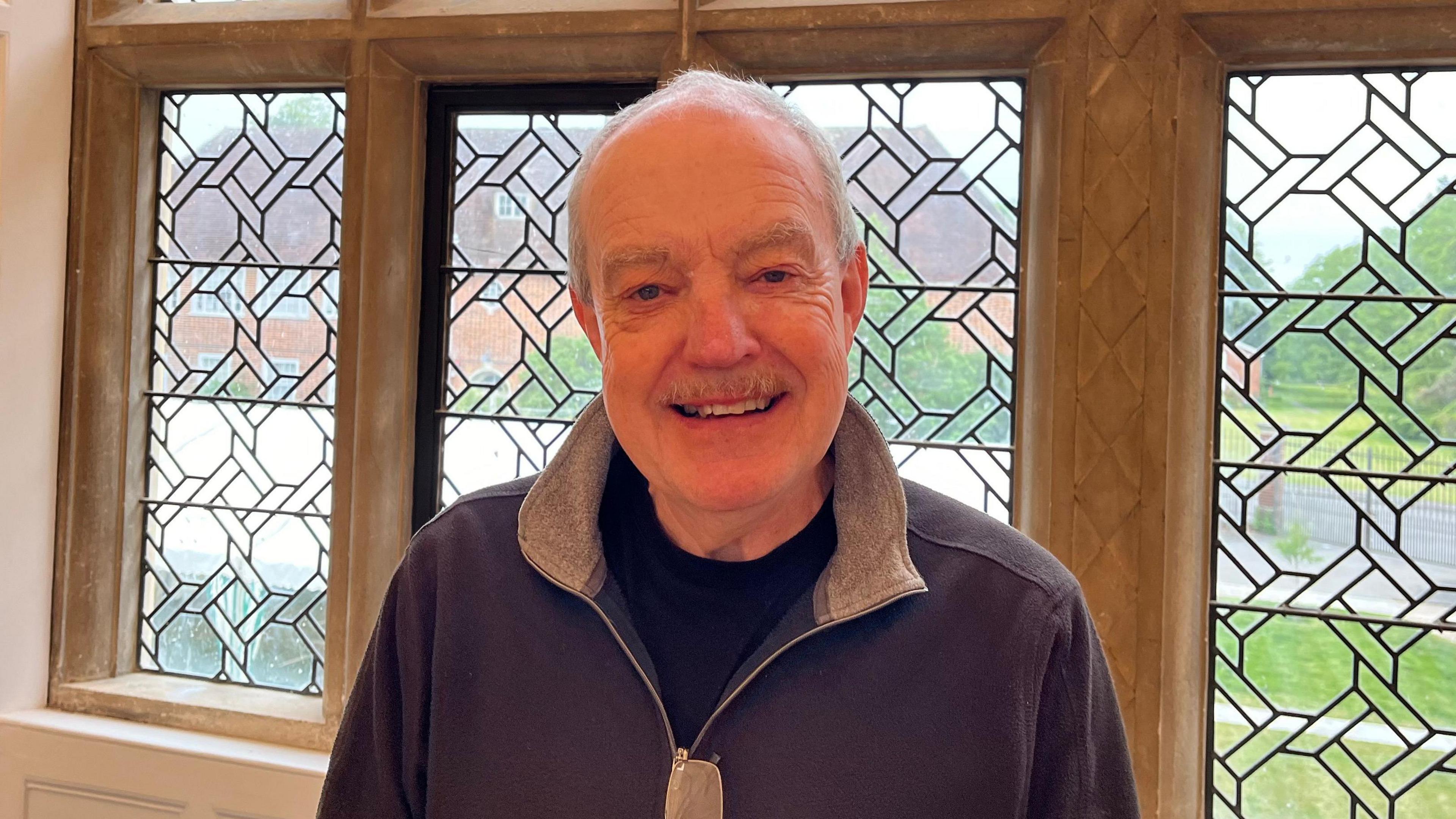 Ken Smith - wearing a blue jumper, with grey hair and moustache, looks into the camera in front of an old stone-framed window