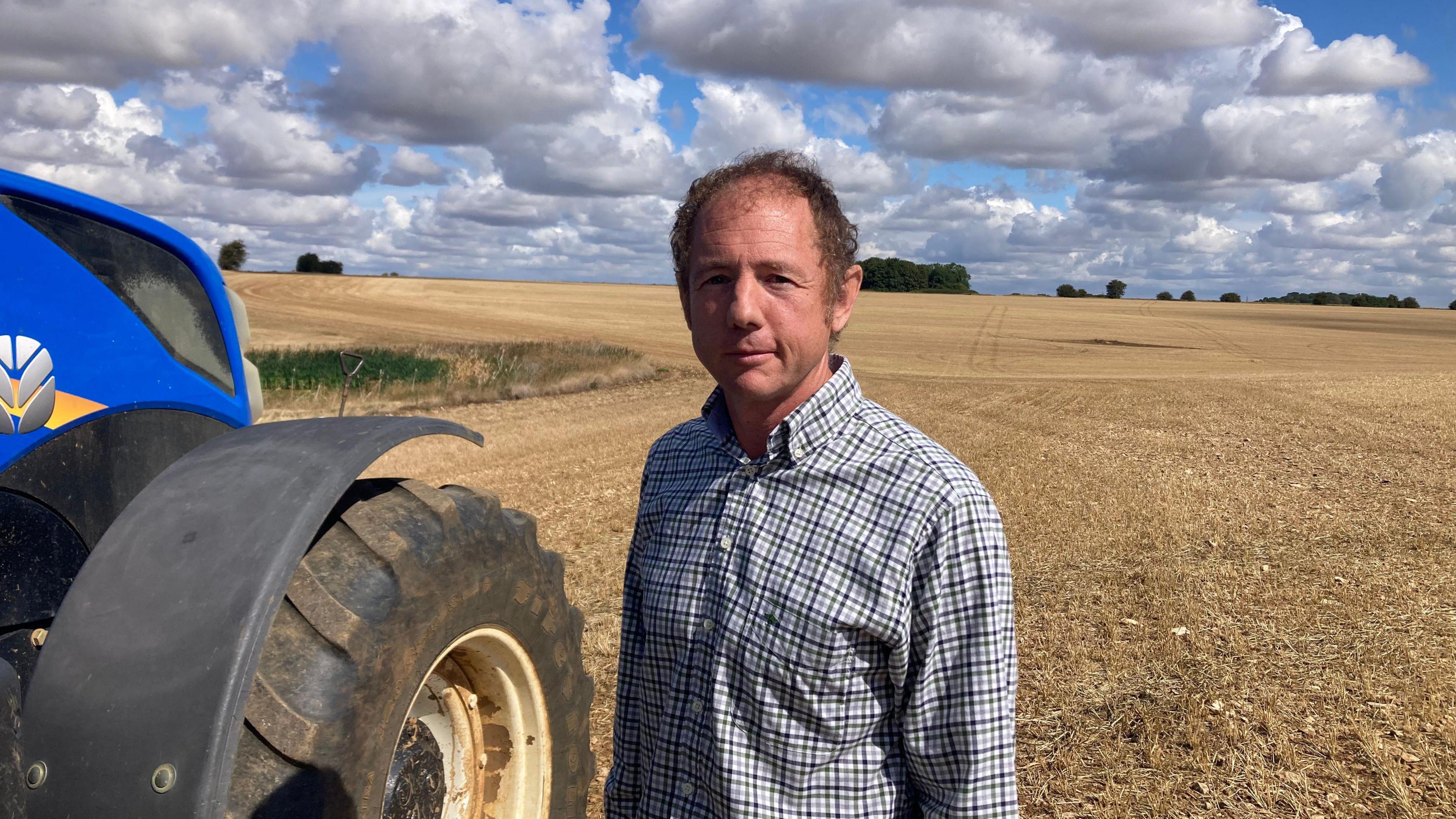 A brown haired man in a chequered shirt stands next to the wheel of a large blue tractor. He's standing on a freshly harvested field in the sun with a bright cloudy sky behind him.