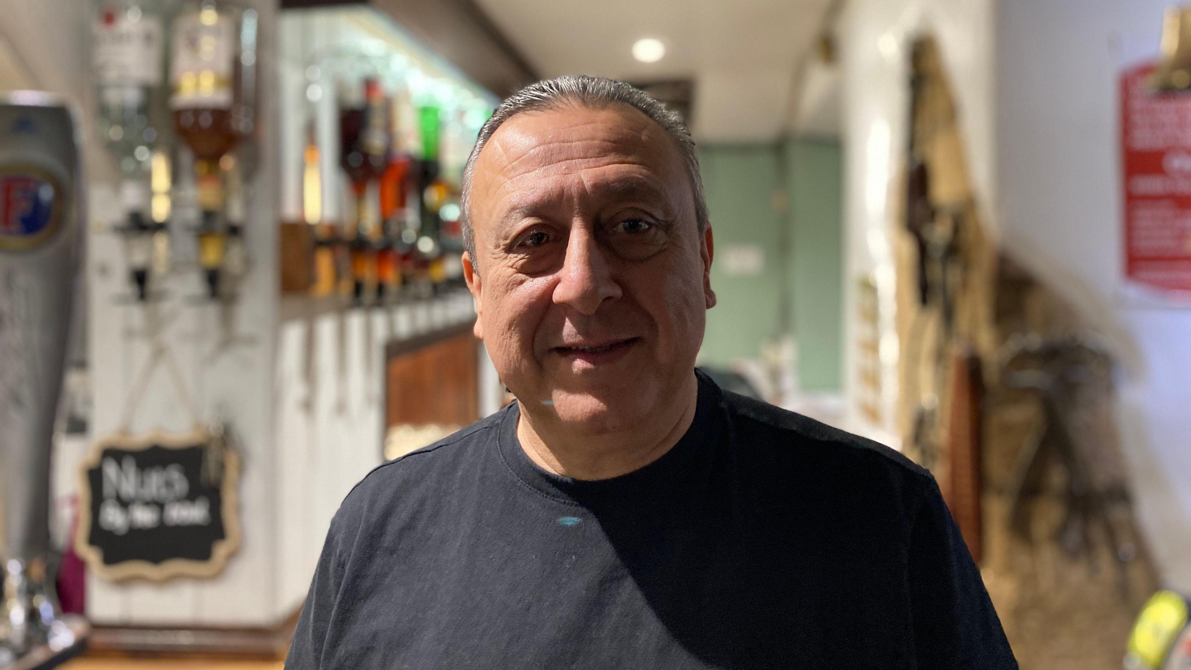 Geoff Sharrif smiles and looks at the camera. He is wearing a black jumper and a pub's bar is behind him. 