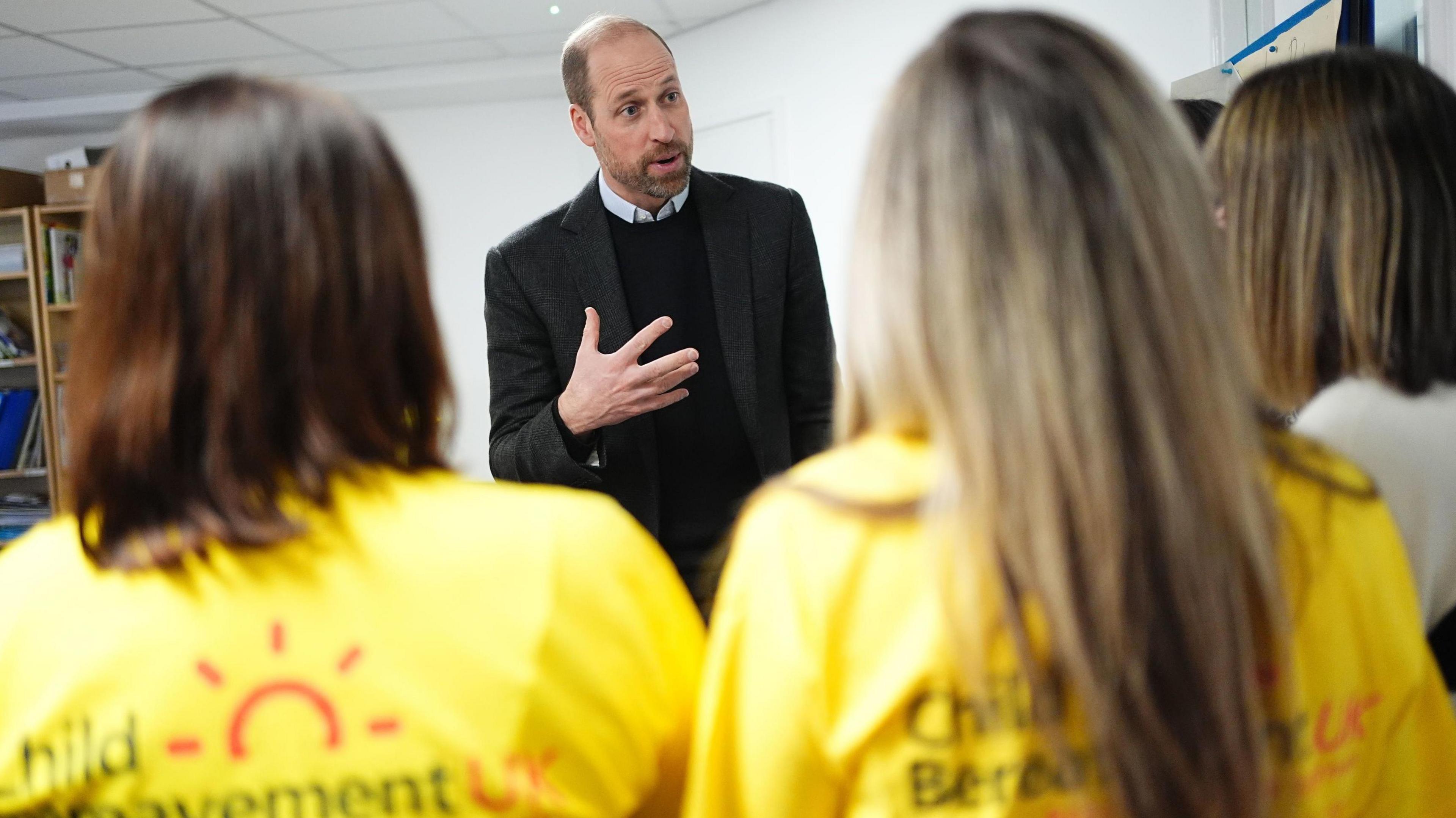 Prince William talks to a group of people. Two women  in yellow charity shirts have their back to the camera.