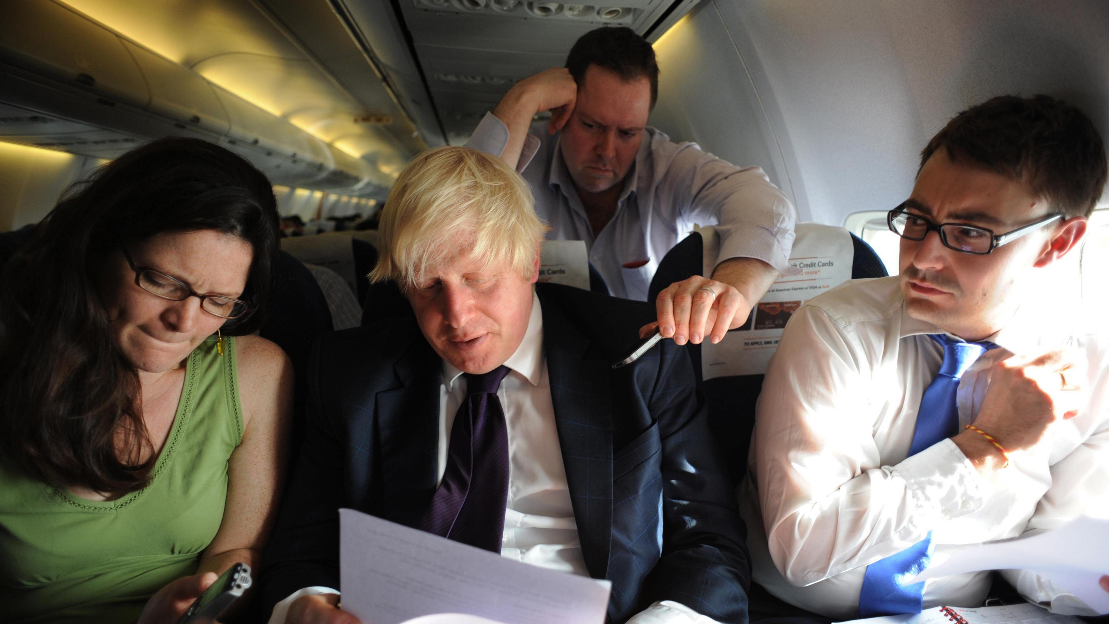 Former UK prime minister Boris Johnson sitting with two people on a plane. Johnson is reading papers. There is a man leaning over the seat.