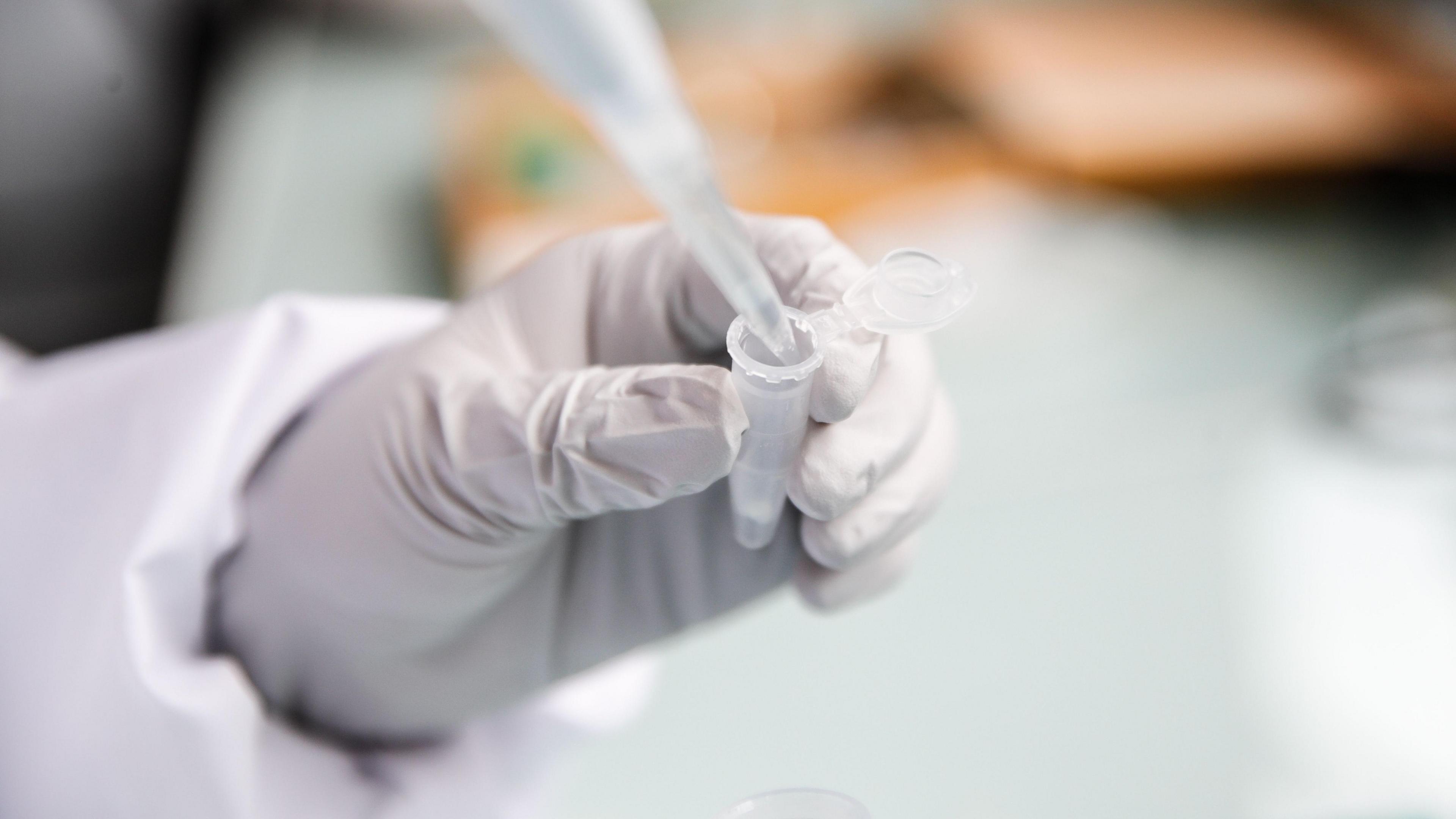 A person wearing rubber gloves and personal protective equipment uses a pipette.