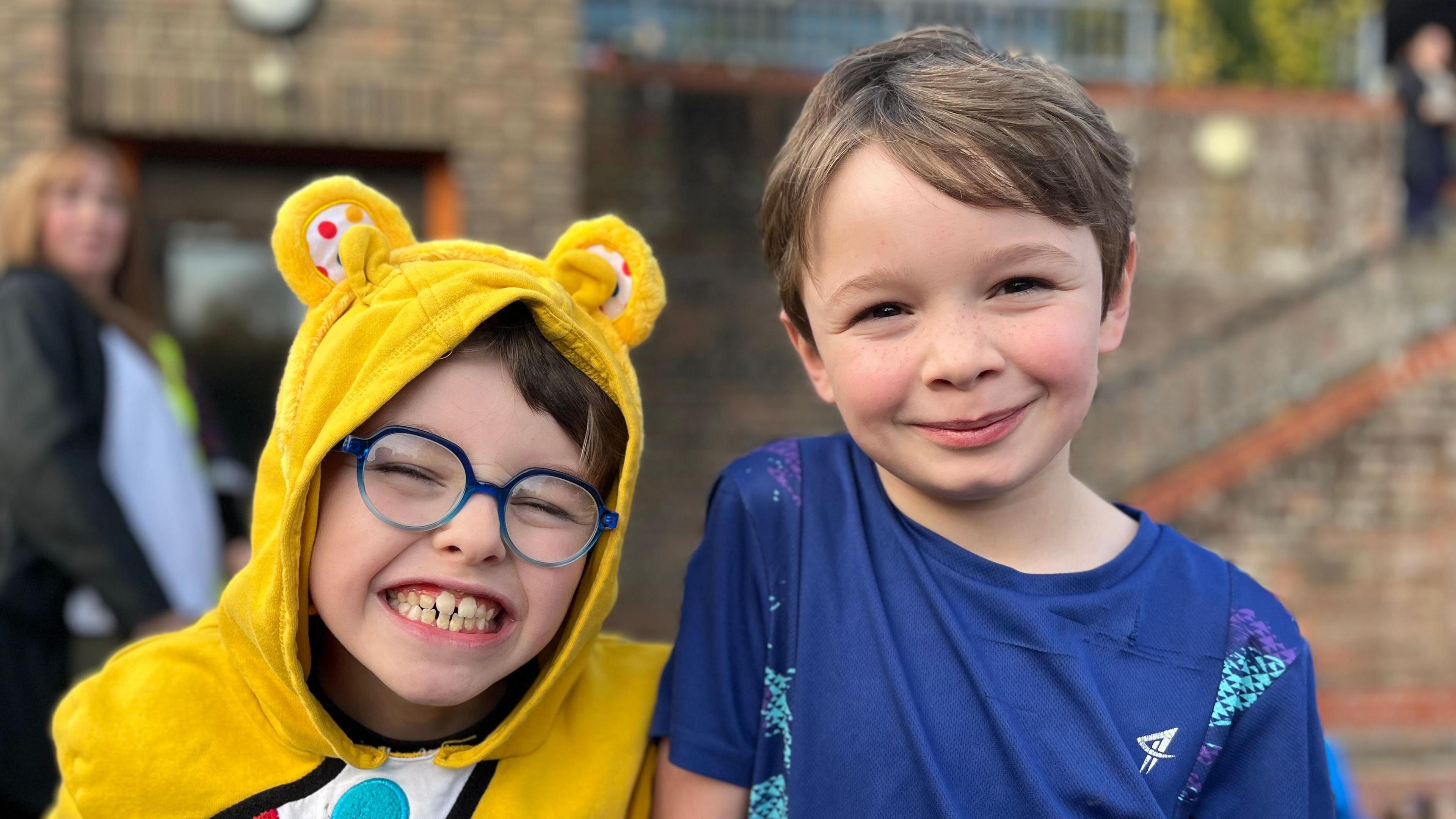 An eight-year-old boy with glasses wearing a Pudsey outfit smiling at the camera. He is standing next to a taller boy his age with a blue running shirt on.