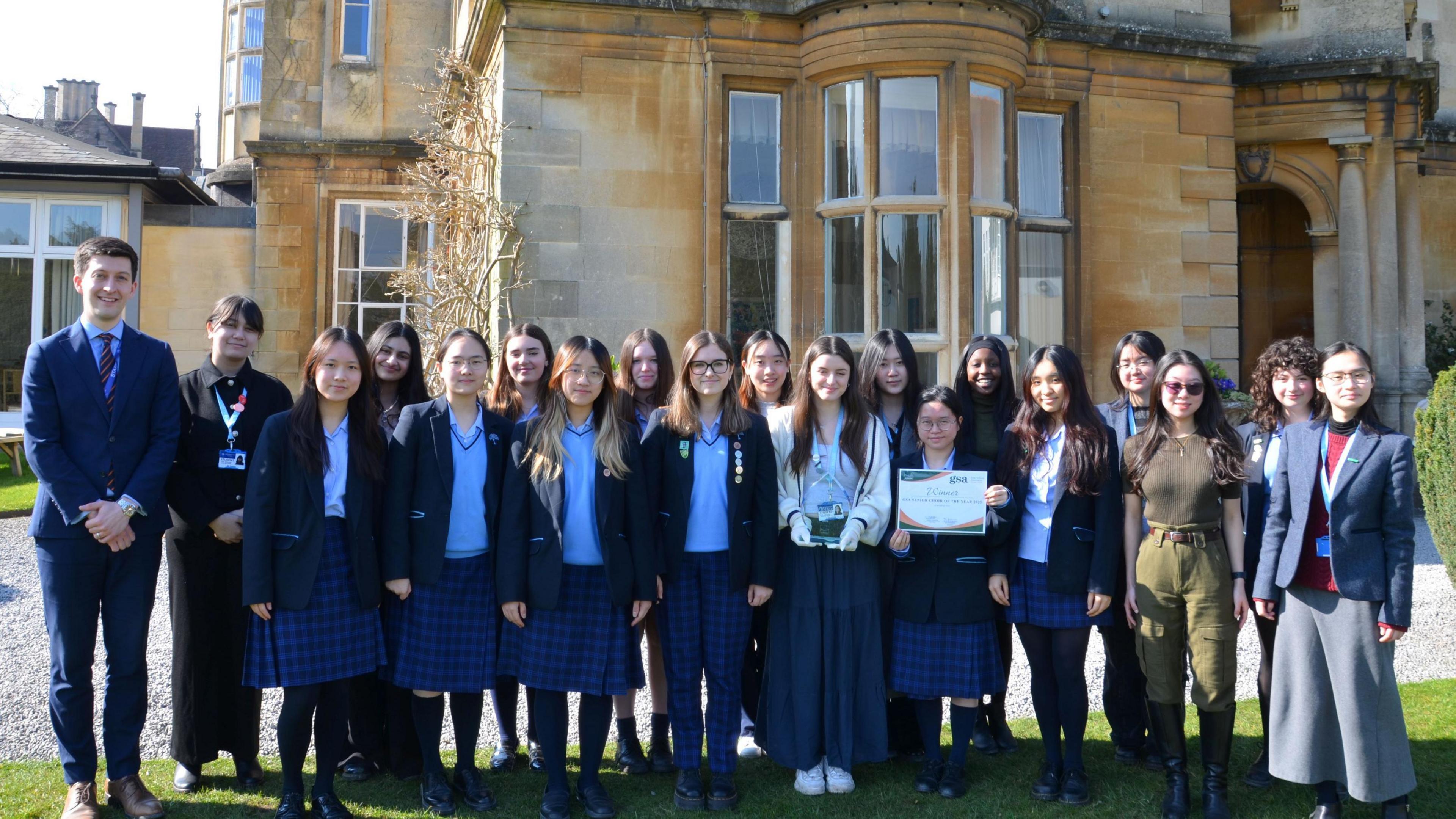 A large group of pupils from Badminton School in Bristol line up to face the camera standing outside the school. They are almost all in the school's uniform of dark blue skirts or trousers and light blue polo tops. In the background is the stone exterior of the school and the photograph is taken on a sunny day