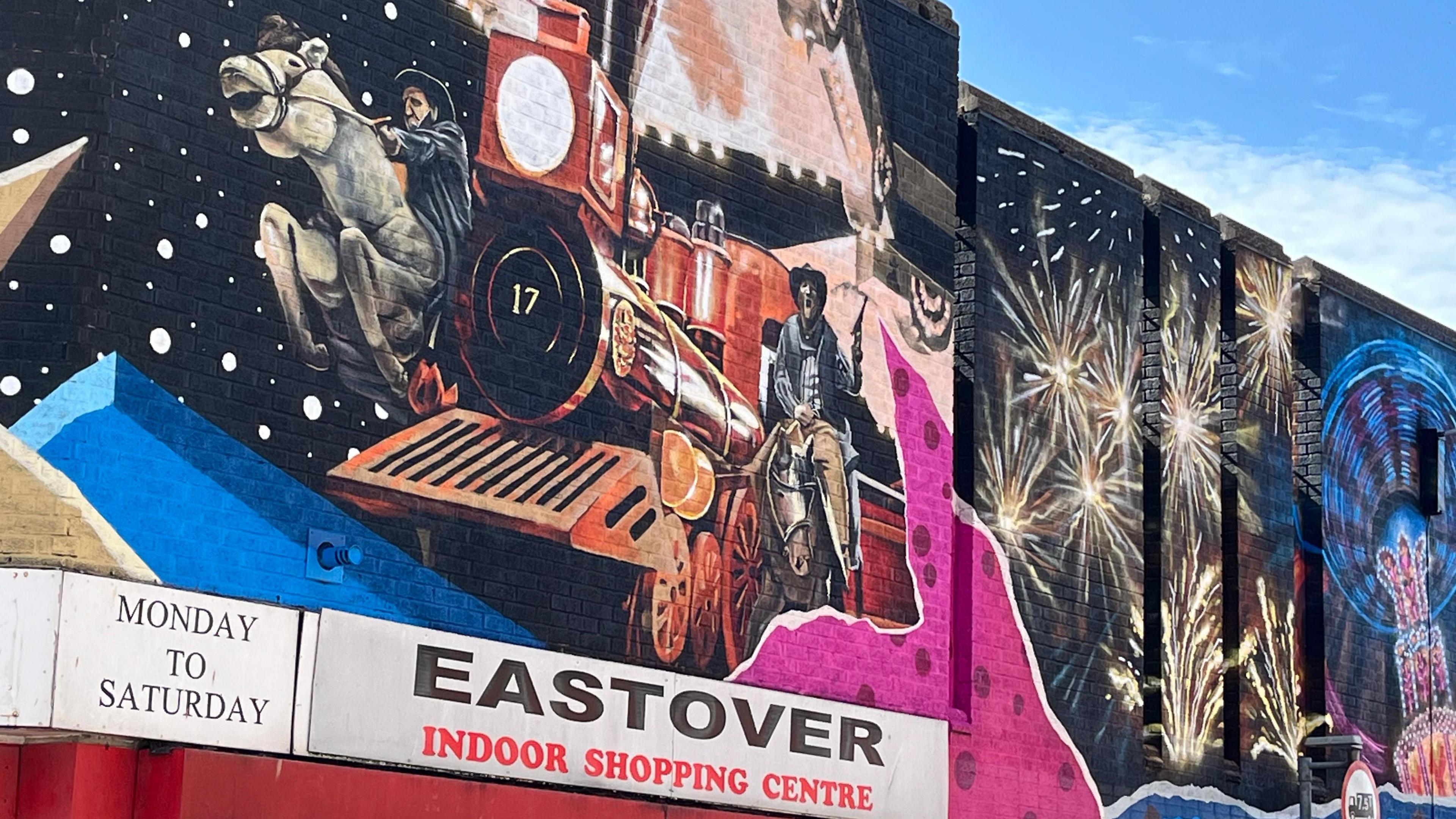 A colourful mural painted on a black background on a wall above the entrance to Eastover Indoor Shopping Centre. The mural includes a red train and two cowboys on horses as well as fireworks and an abstract illuminated fairground ride