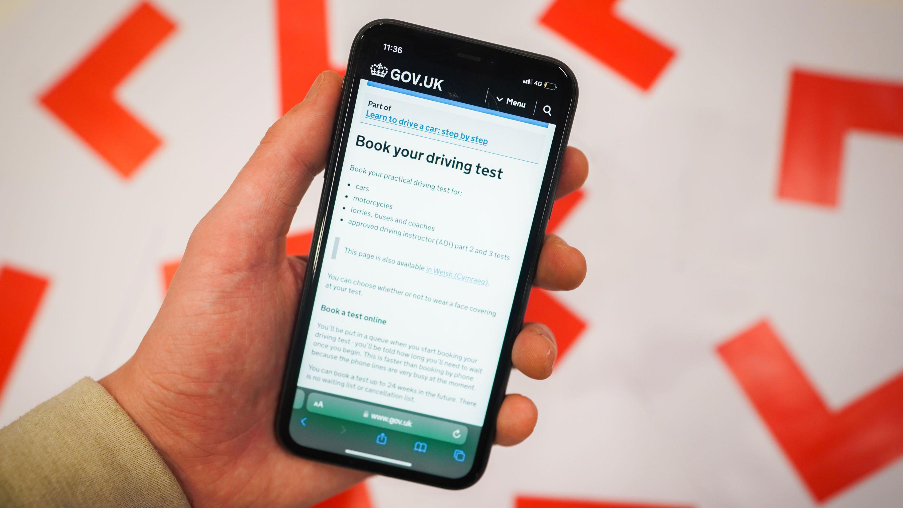 An iPhone held against a backdrop of L plates. On the phone is the website to book a driving test.