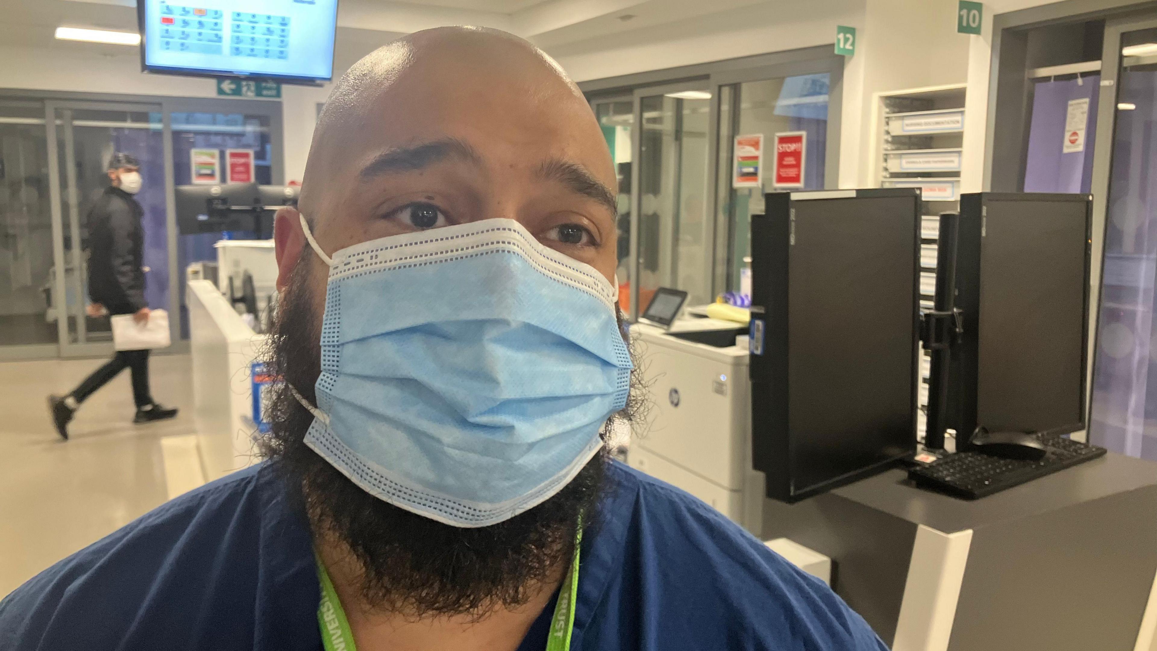 A doctor in scrubs and a face mask standing in a busy hospital department