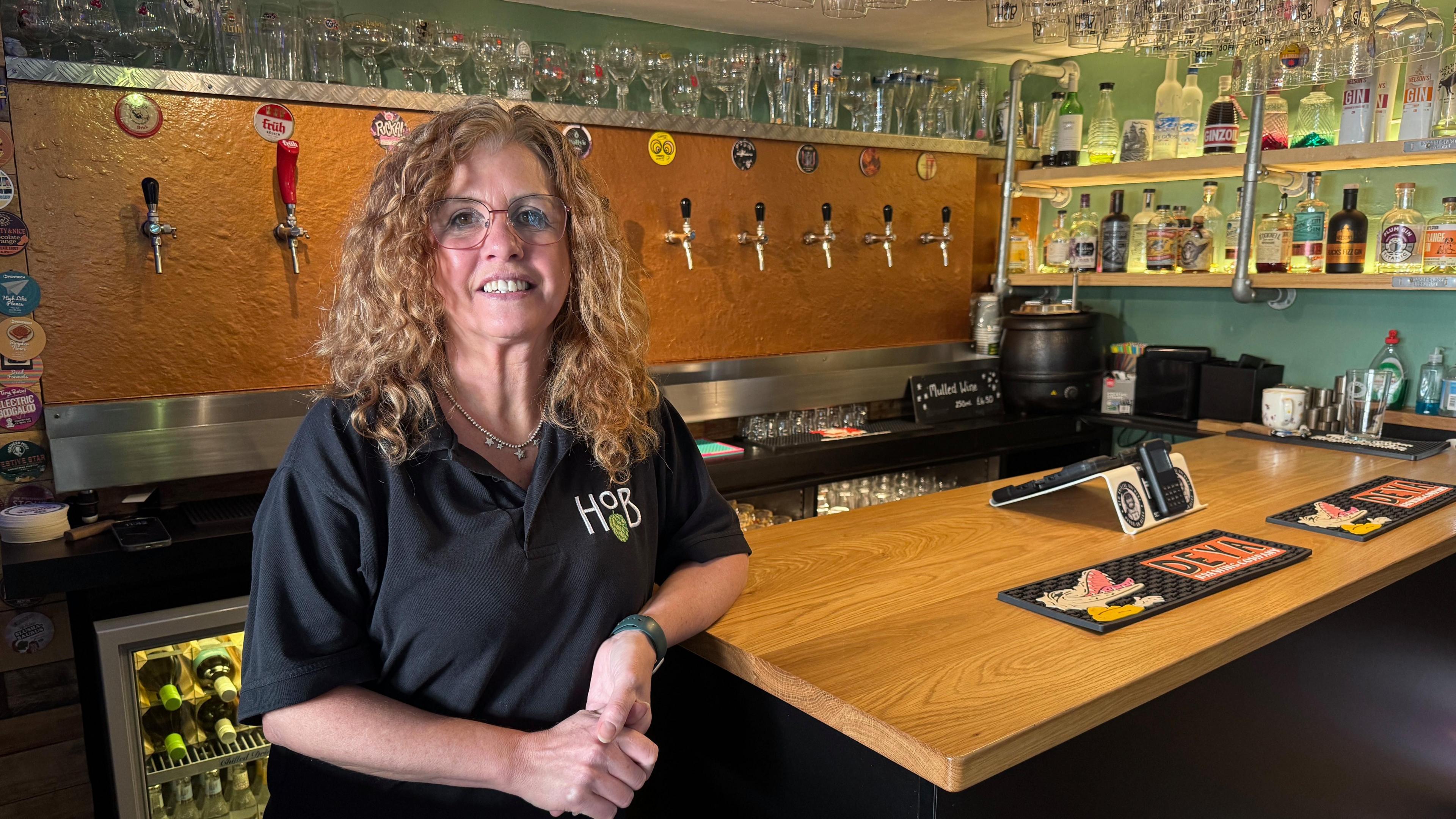 Amanda Hodson Walker (long curly hair, glasses)  wearing a black logoed t shirt leaning on the corner of the bar with a row of modern taps on the back wall, drinking glasses above and spirits on the right hand wall.