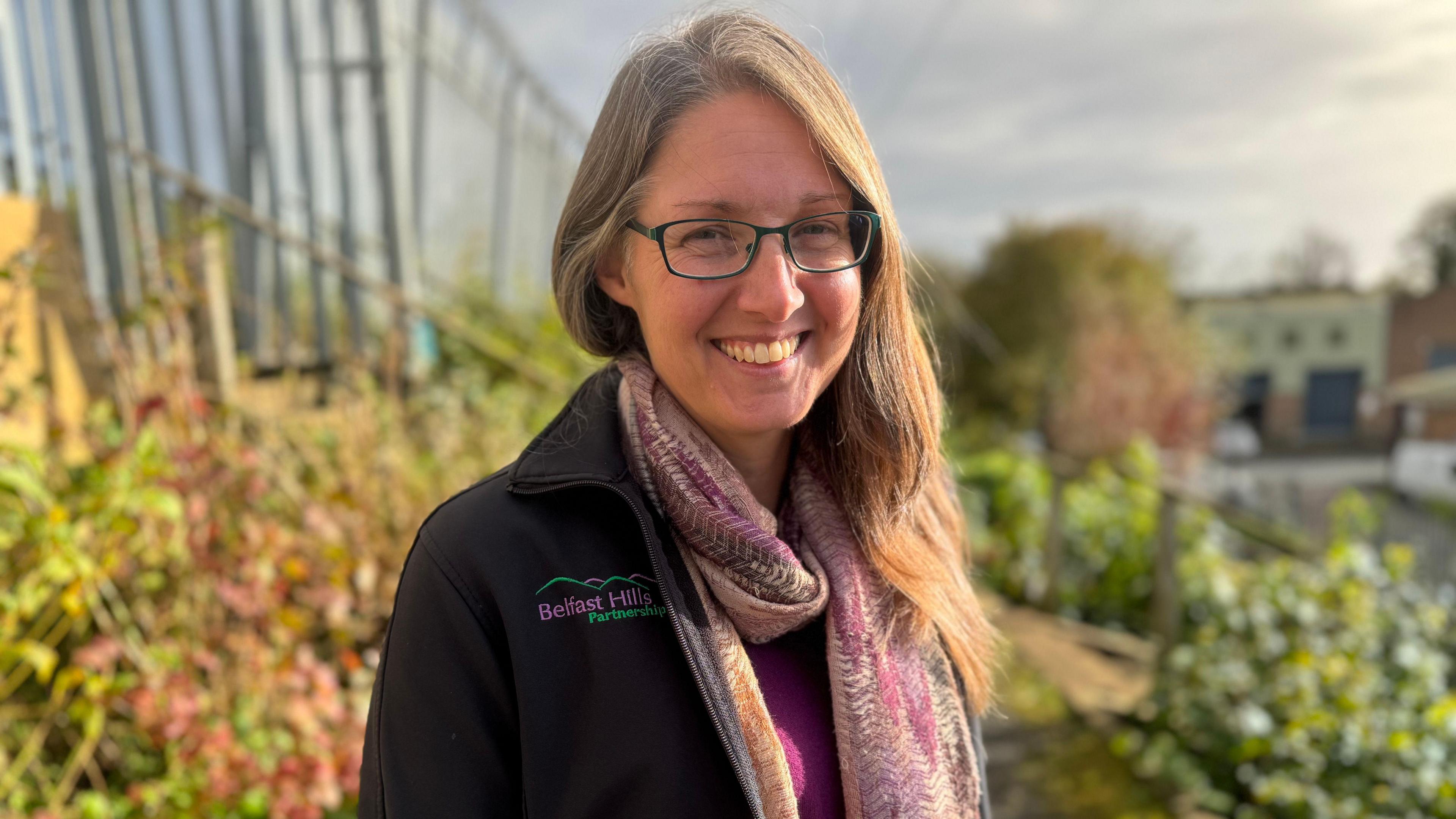 Dr. Lizzy Pinkerton wearing a purple and orange scarf and a black coat standing amongst rows of plants. She has long blonde hair and is wearing glasses with rectangular black rims.