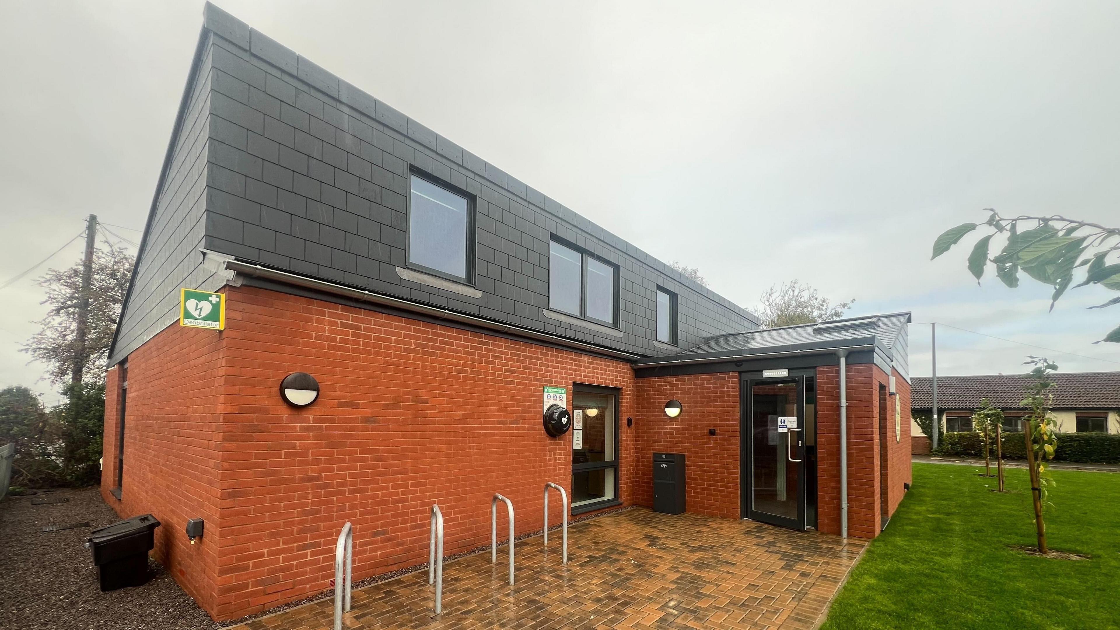A two storey community building with silver bicycle racks outside. The first storey is red brick and the top is slate tiles. It is fronted by green lawn,