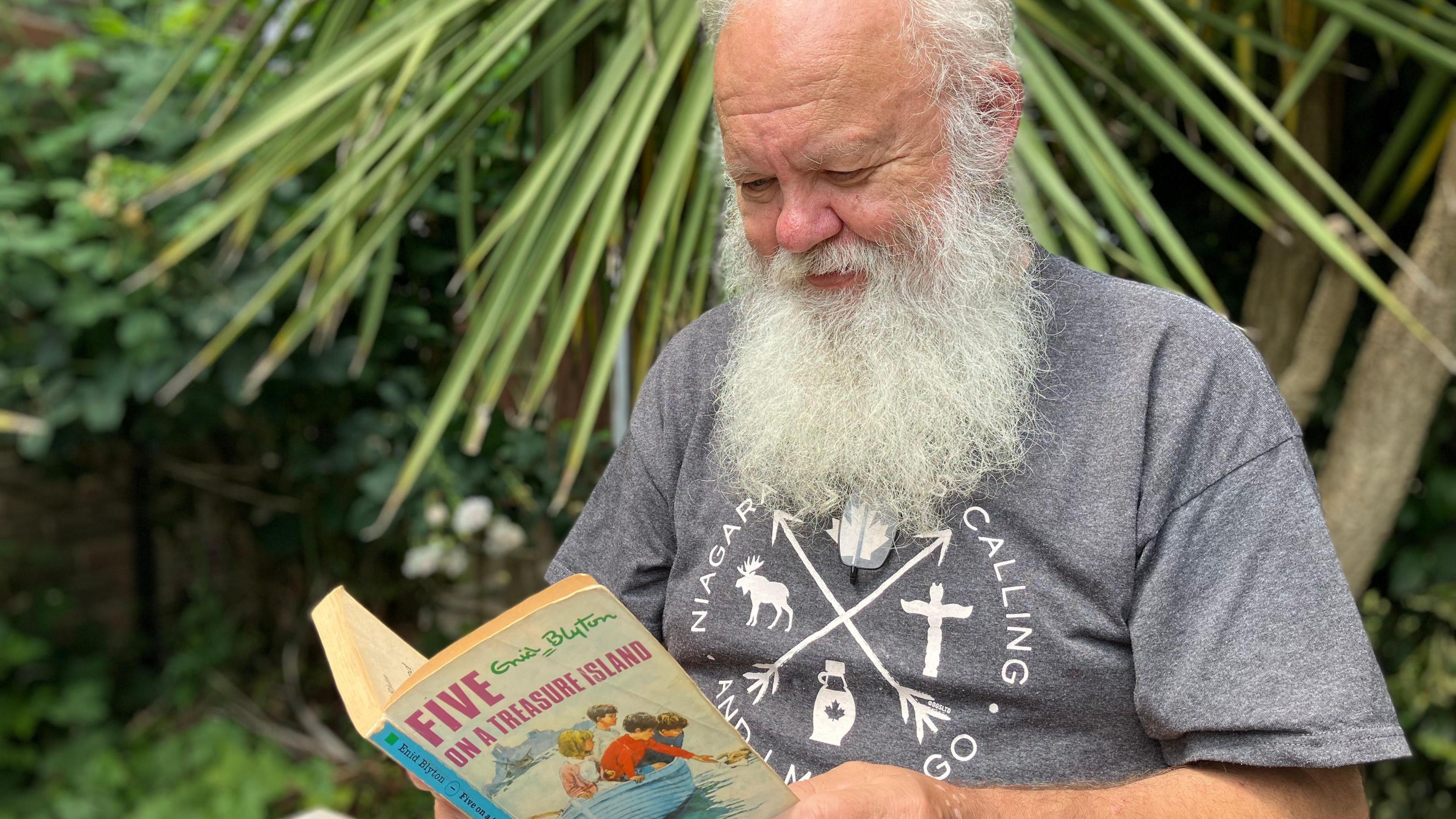 Mr Mills reads a book in front of plants and flowers in his front garden. He looks focused but is smiling at the same time.