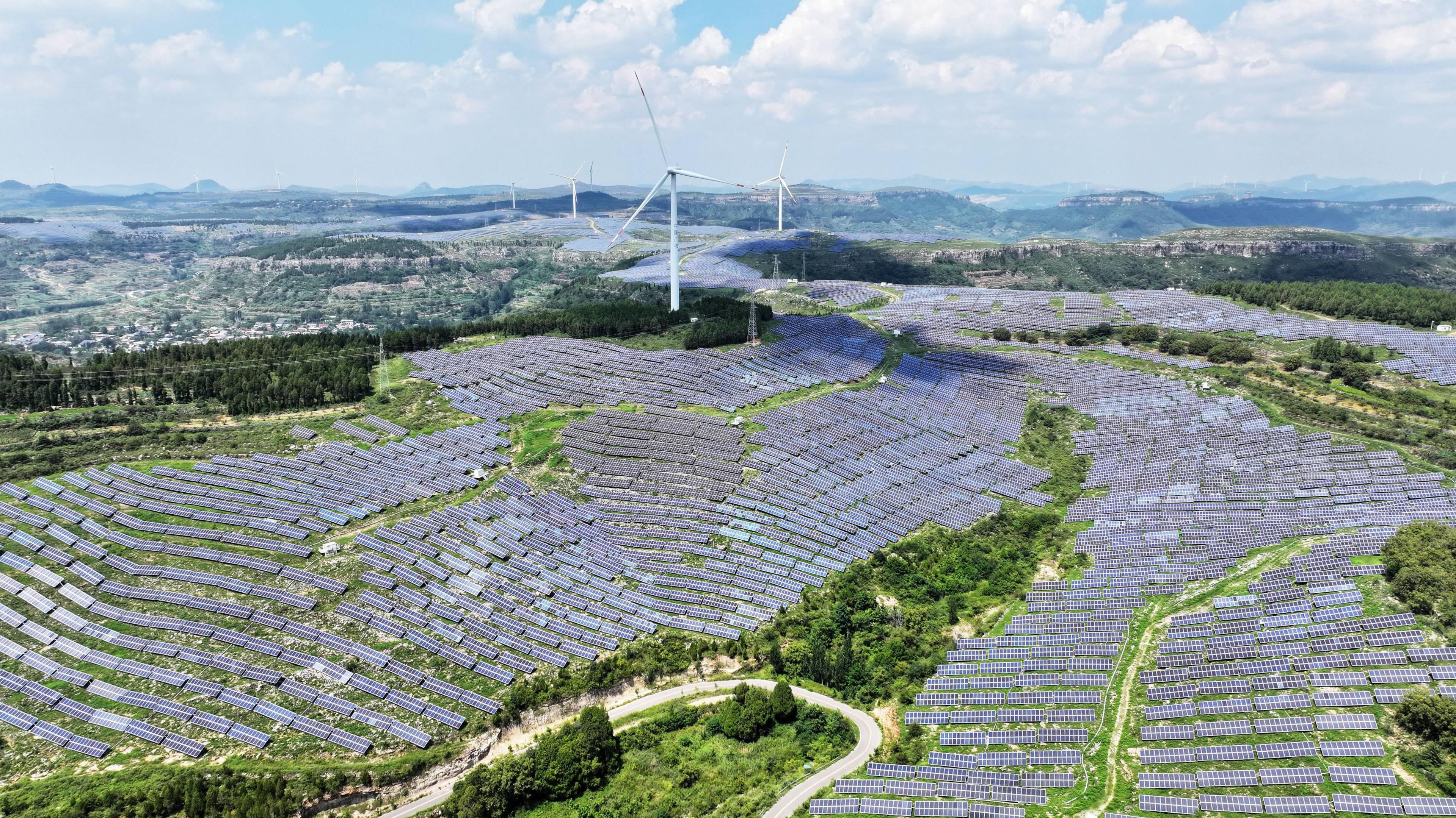 An aerial view of a photovoltaic solar farm sprawling across undulating green hills in Shandong Province, China. The are also several white wind turbines rising in the background. A winding road curves through the lower foreground.