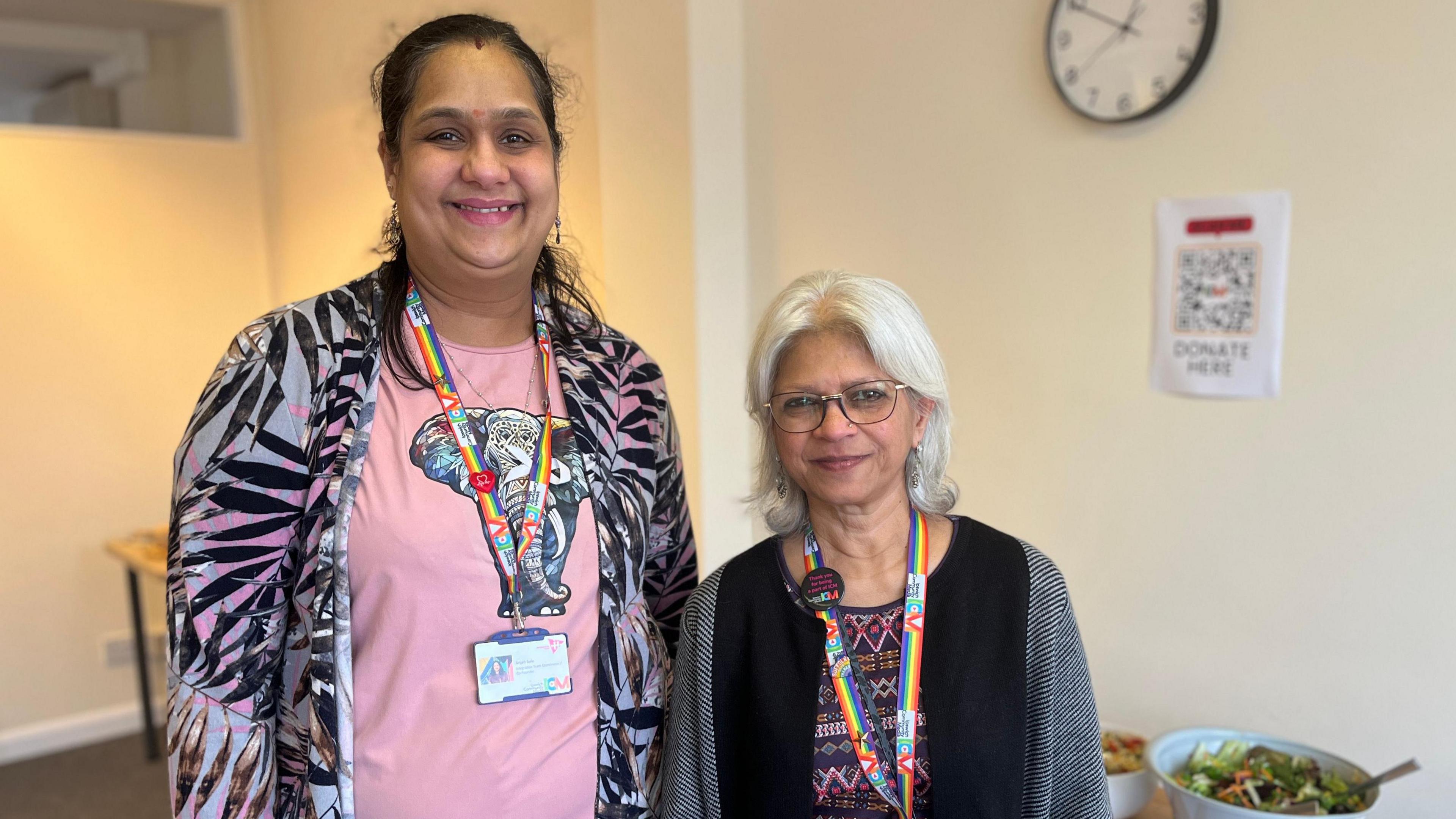 Anjali Sule and Pratchi Katdare in a new, large room, with food on a table behind them. Anjali is wearing a pink t-shirt with an elephant illustration on it, and a palm leaf printed cardigan. Pratchi has white hair and is wearing a black and grey cardigan. Both women appear to be of South Asian heritage.