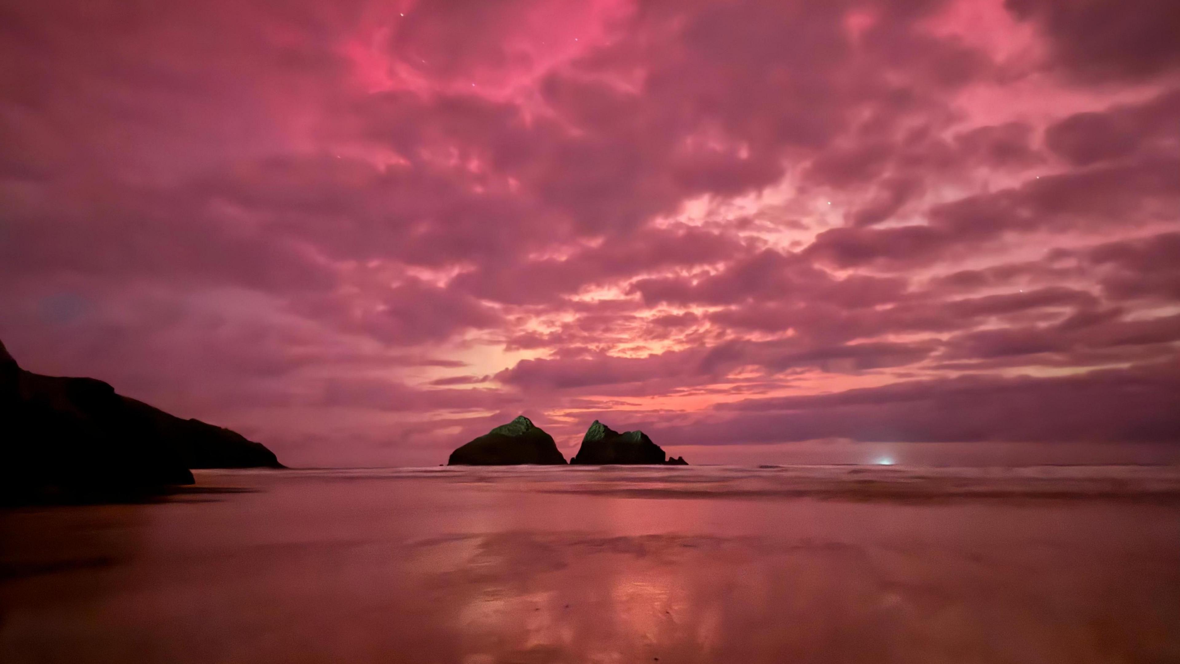 The pink sky shines over Holywell Bay. Two large rocks are visible to the centre of the screen.