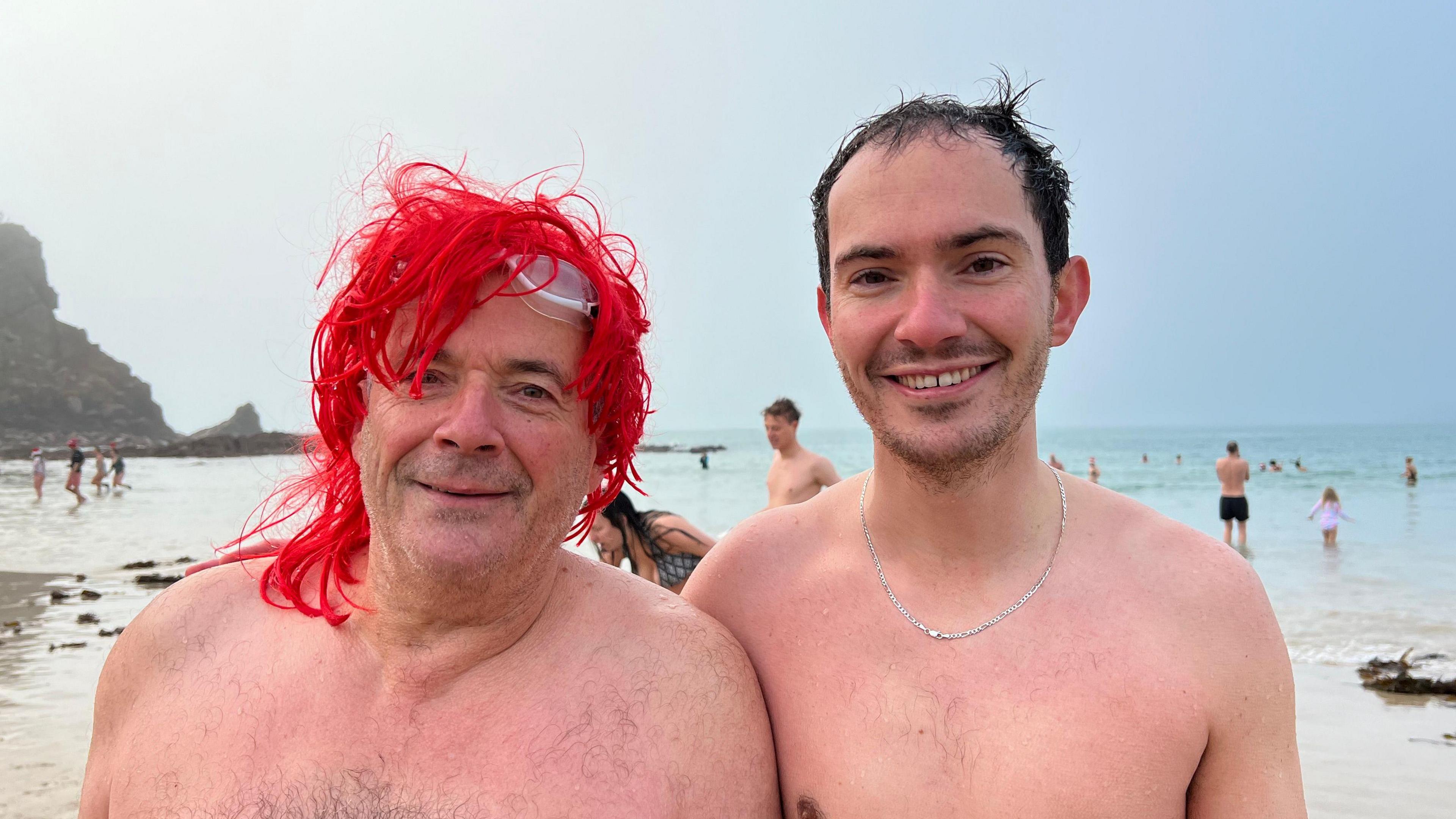 Martin Tupper is a middle-aged man with no shirt on, wearing a red wig. Max is a younger man with dark hair, with no shirt on and a silver chain. They are standing on the beach with swimmers in the sea behind them.