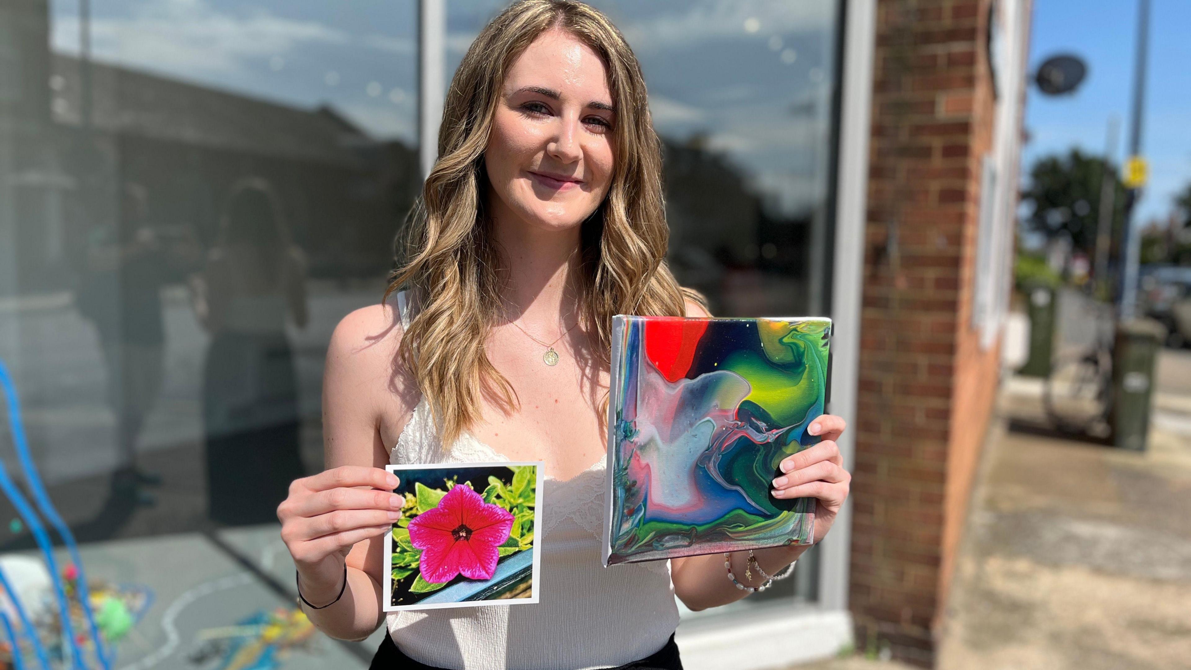 A woman holds two images of flowers. She is standing in front of a gallery window.