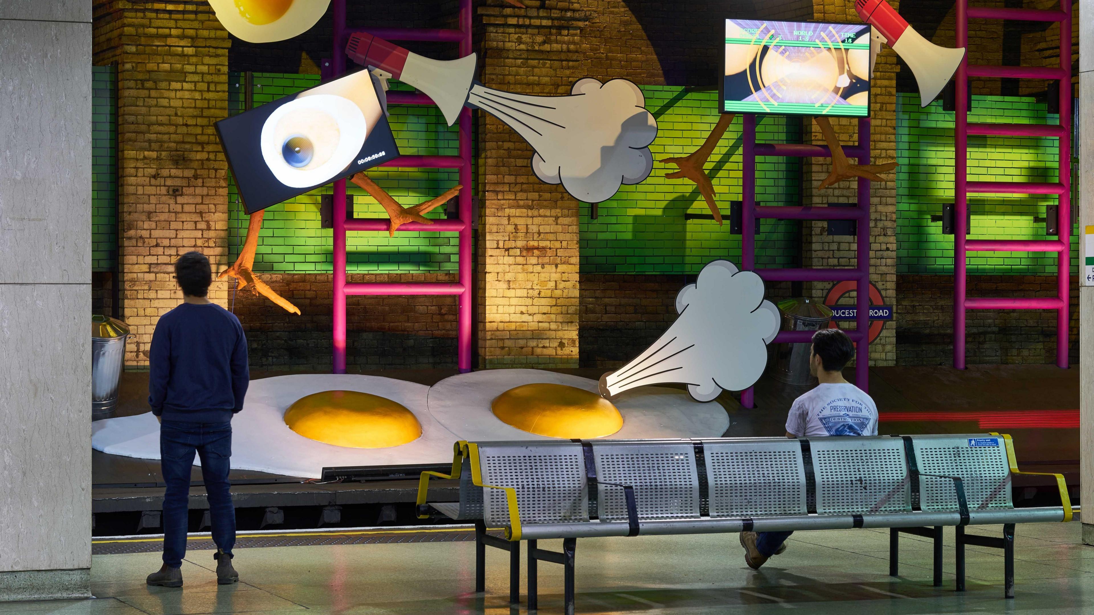 Two people with their backs to the camera, one sitting on a bench and one standing, look across the platform to a surreal art exhibit featuring two giant cardboard cracked eggs with yellow yolks, there are ladders, megaphones and signs indicating wind coming out of the eggs and the megaphones. 