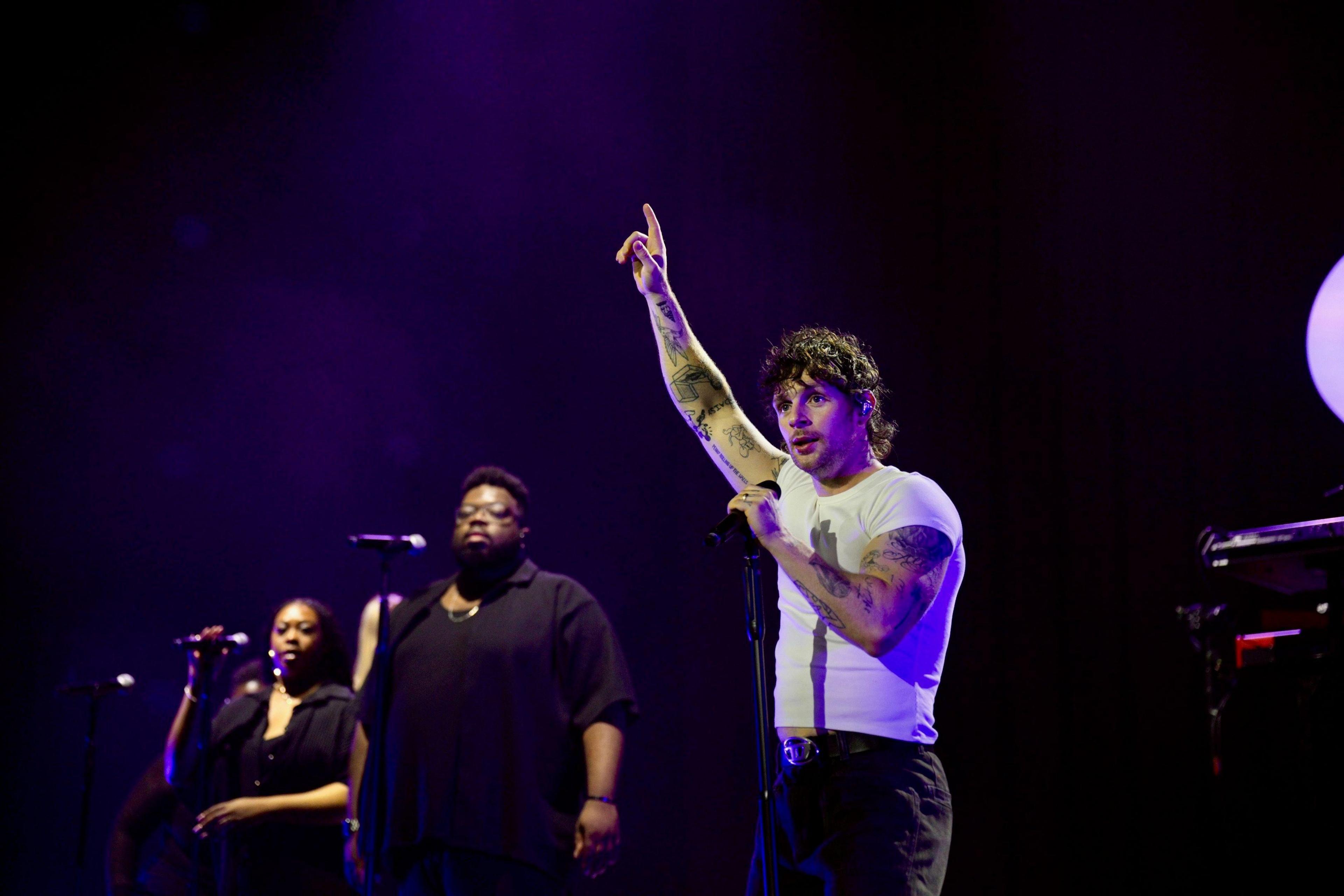 Singer Tom Grennan is on stage wearing a white T-shirt and black trousers, He has one arm raised and two singers wearing black outfits are on his right.