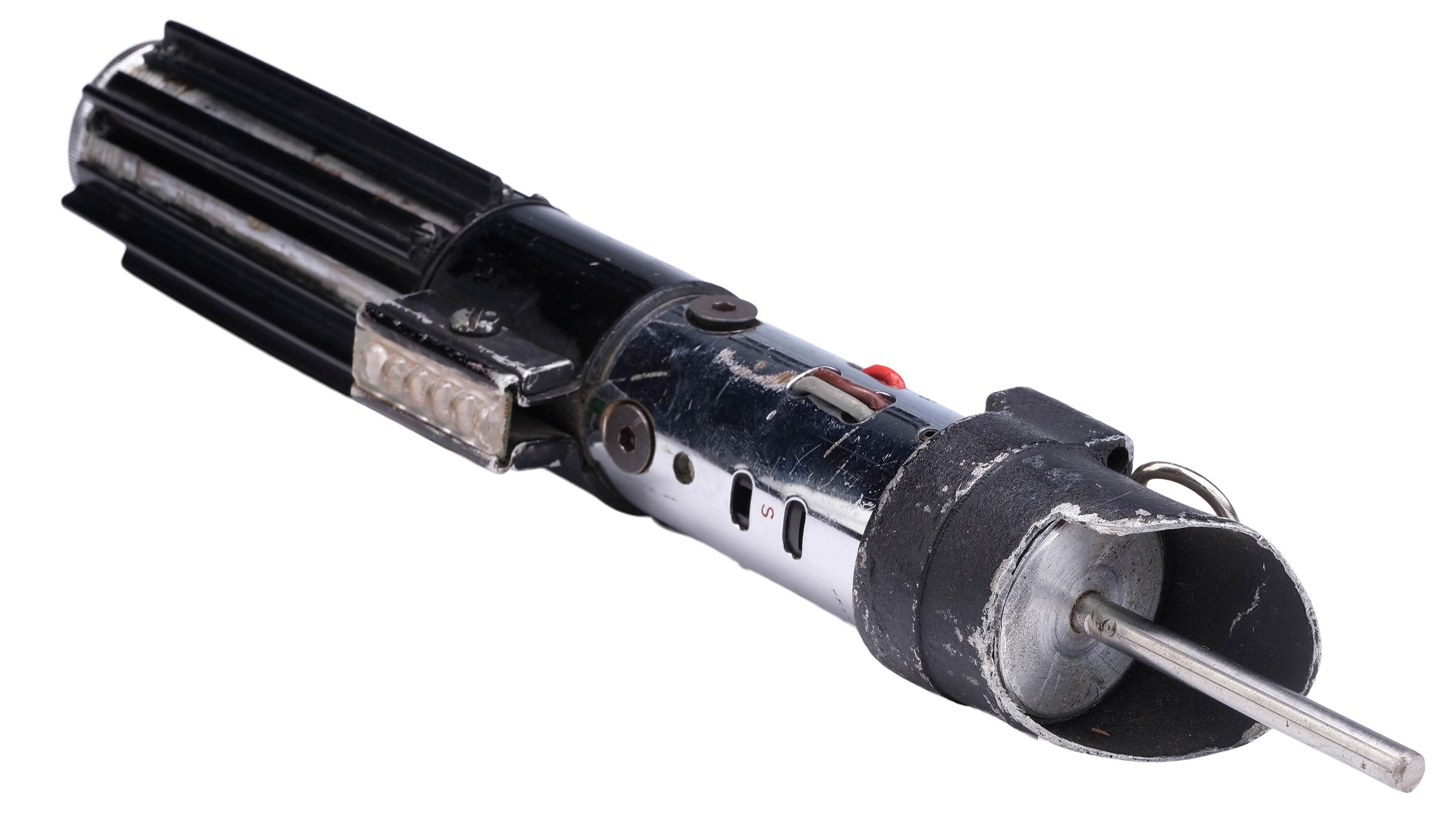 Darth Vader's lightsaber, lying on its side against a white backdrop. A metal prong protrudes from the top, where a blade would have been attached.