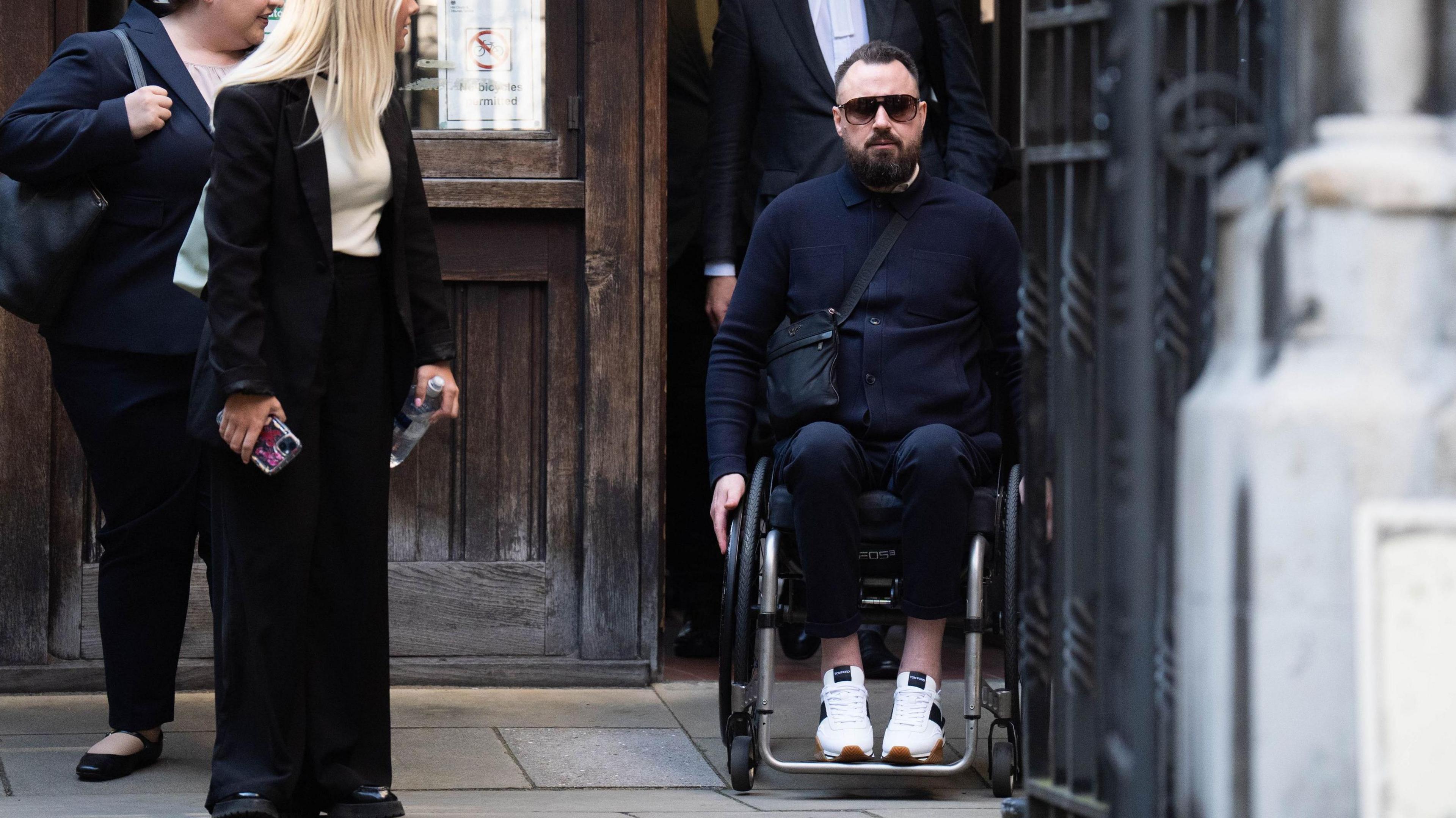 Martin Hibbert, who was injured in the May 2017 Manchester Arena bombing, outside the Royal Courts Of Justice, central London. He is wearing a navy blue top with long sleeves, dark trousers and white trainers. He wears dark sunglasses and is sitting in a wheelchairl.