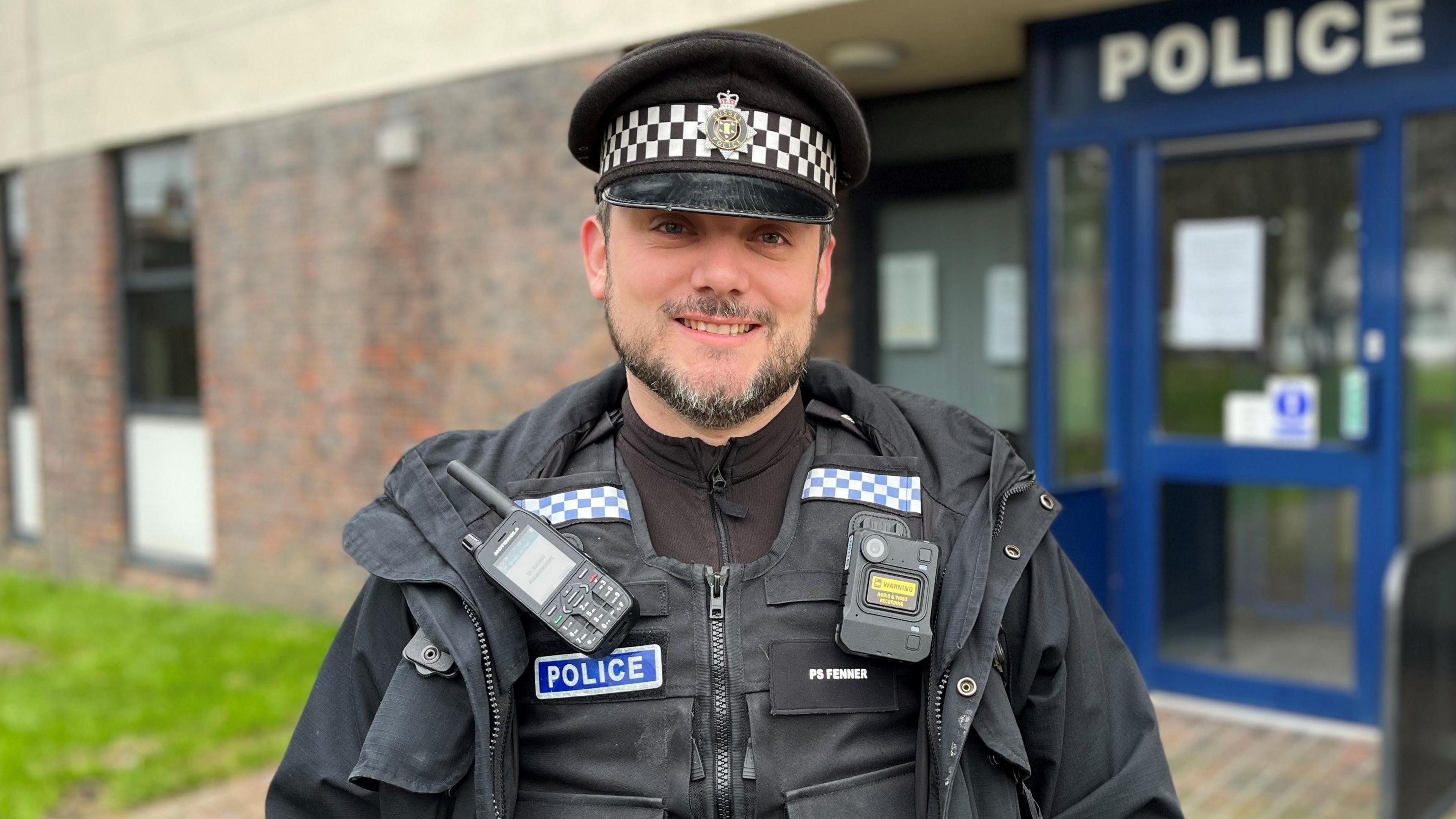 A policeman with a dark beard and a police coat and hat on. He is standing in front of a blue door which has "POLICE" written above it in white letters.