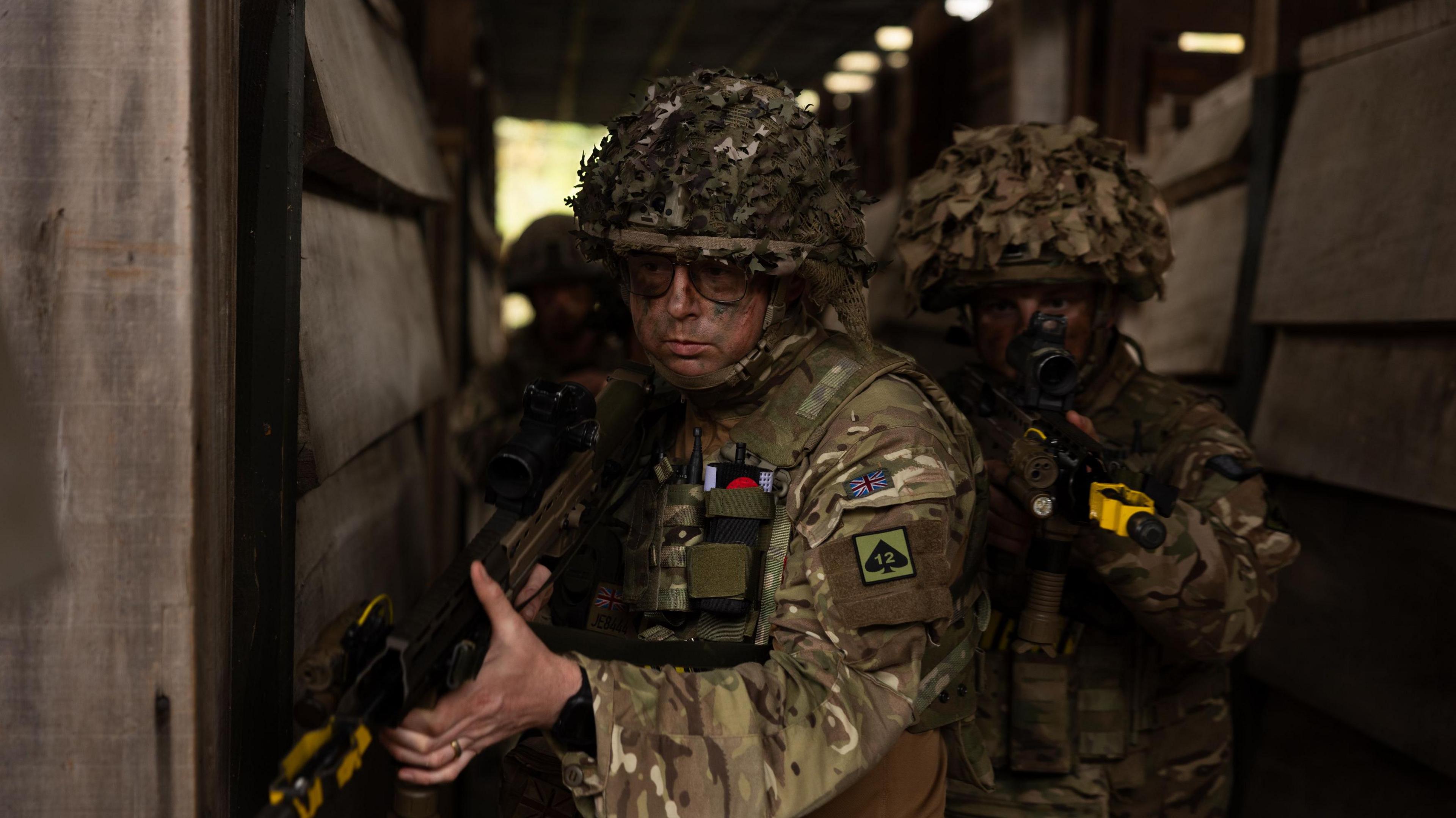 Three men in camouflage holding guns are stood in what looks like an outdoor wooden hallway.