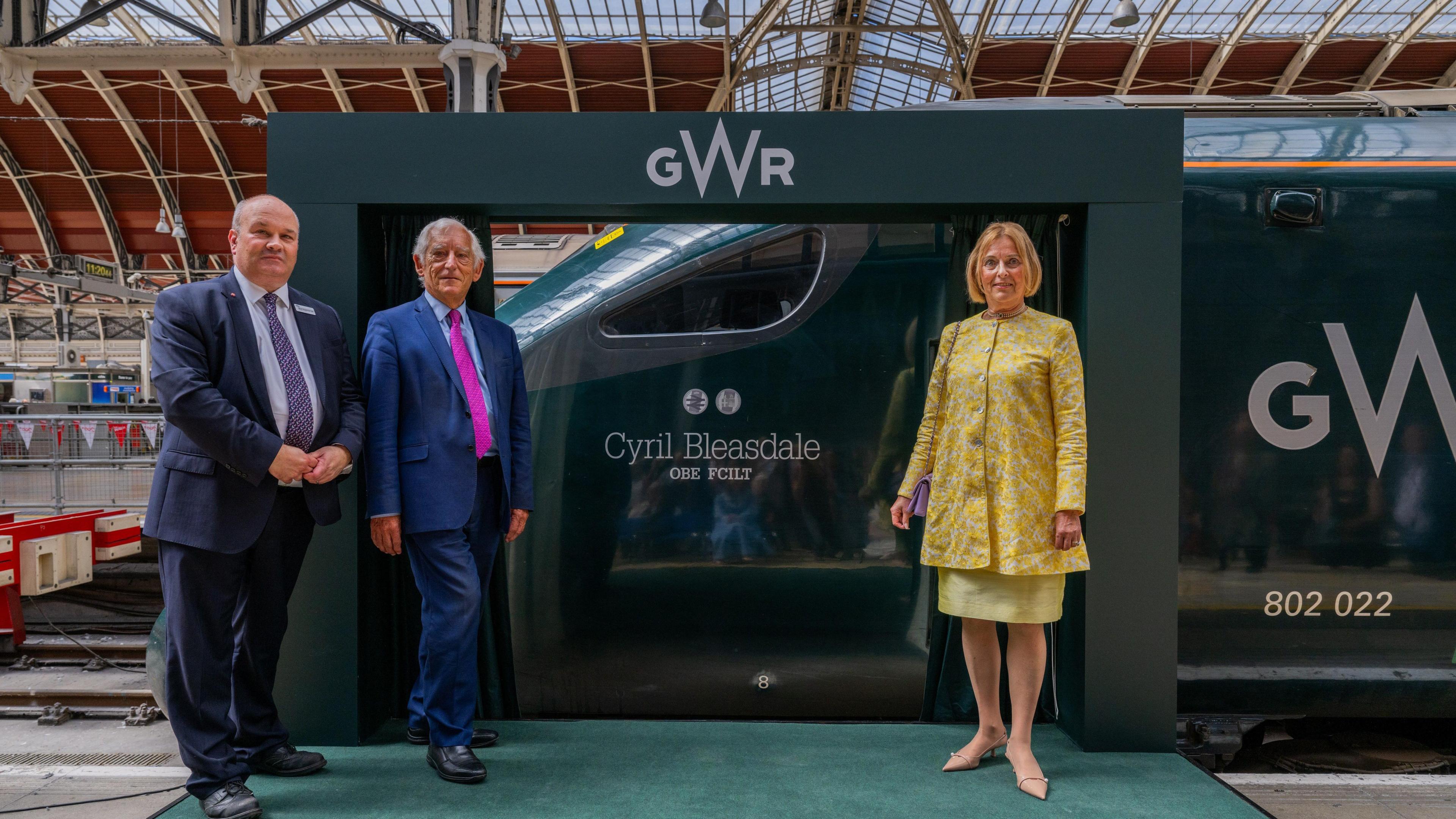 GWR managing director Mark Hopwood and former InterCity managing director Chris Green in navy blue suits with Cyril's widow Katy in a yellow skirt suit on the platform in front of the GWR train named after Cyril Bleasdale OBE.
