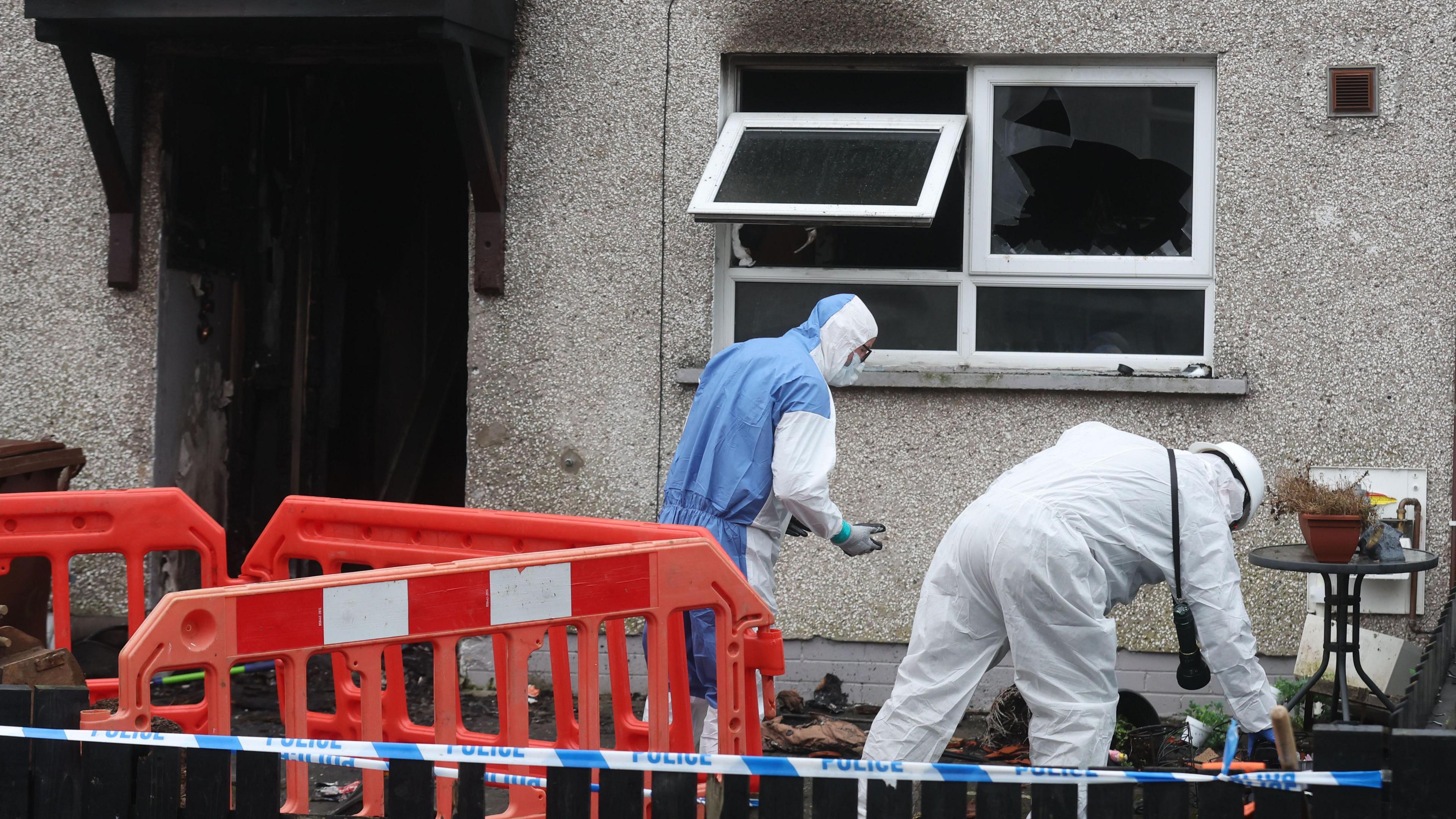 The house is cordoned off with blue police tape and orange safety fences. There are two people in white and blue crime suits lifting rubble. The front of a house with black smoke marks on the doorway and the windows - all windows are open, with one smashed window. There is debris on the floor outside the front of the house and an orange bollard with a forklift. 