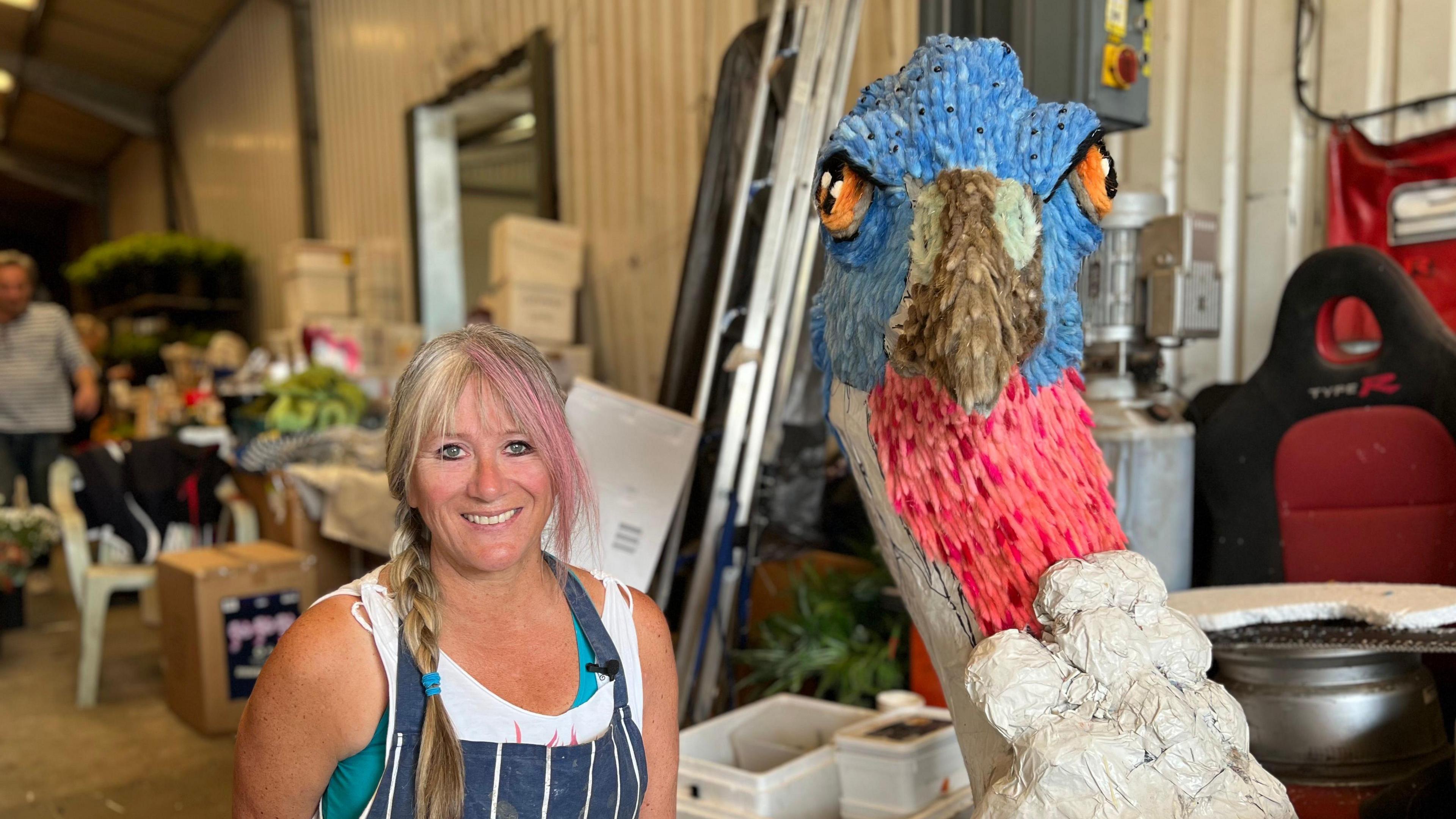 A woman in an apron next to a half-finished turkey head made of papier mâché and blue and pink harestails