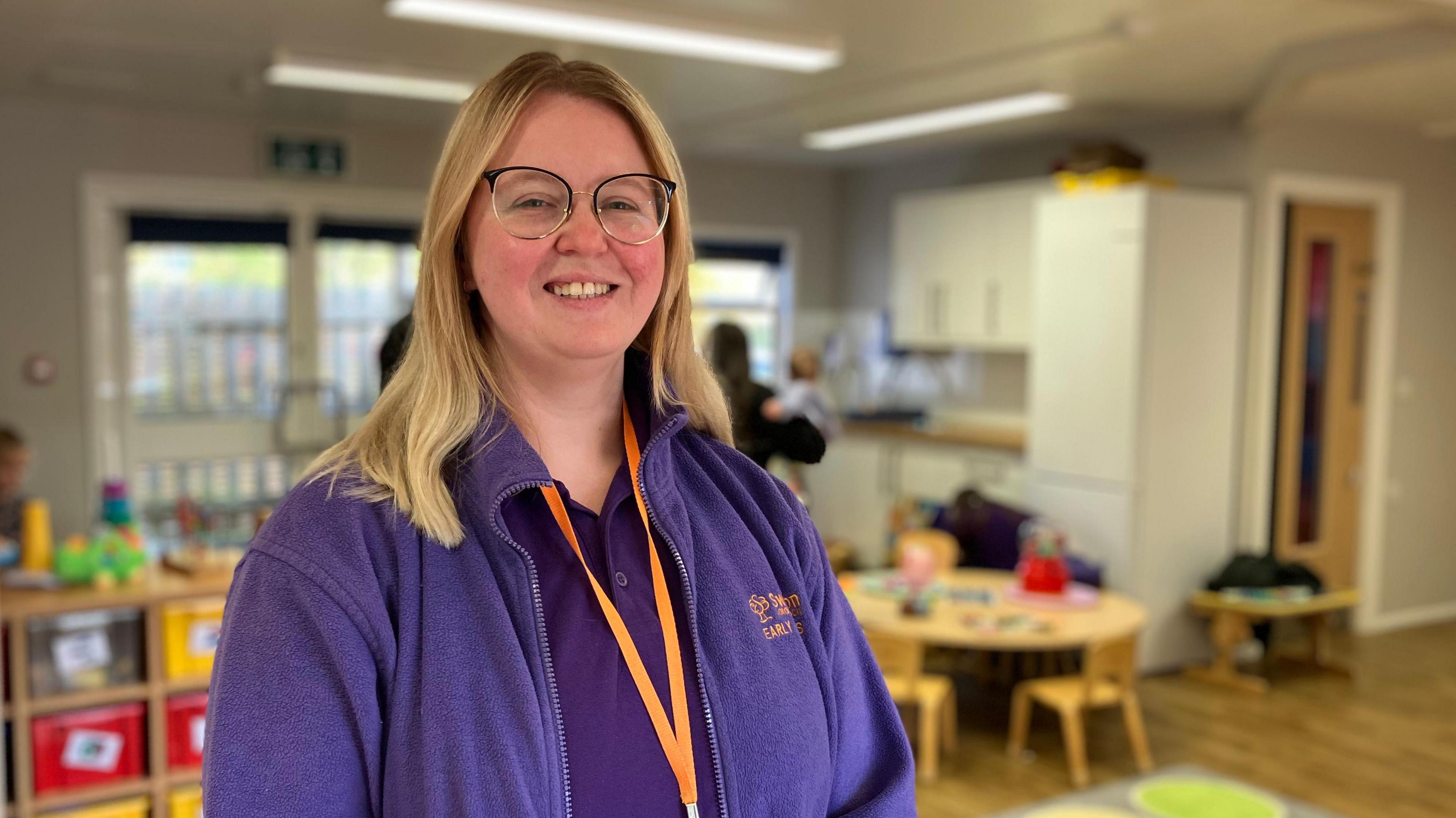 Lauren Love in a purple jumper with an orange lanyard with a blurred school setting behind her