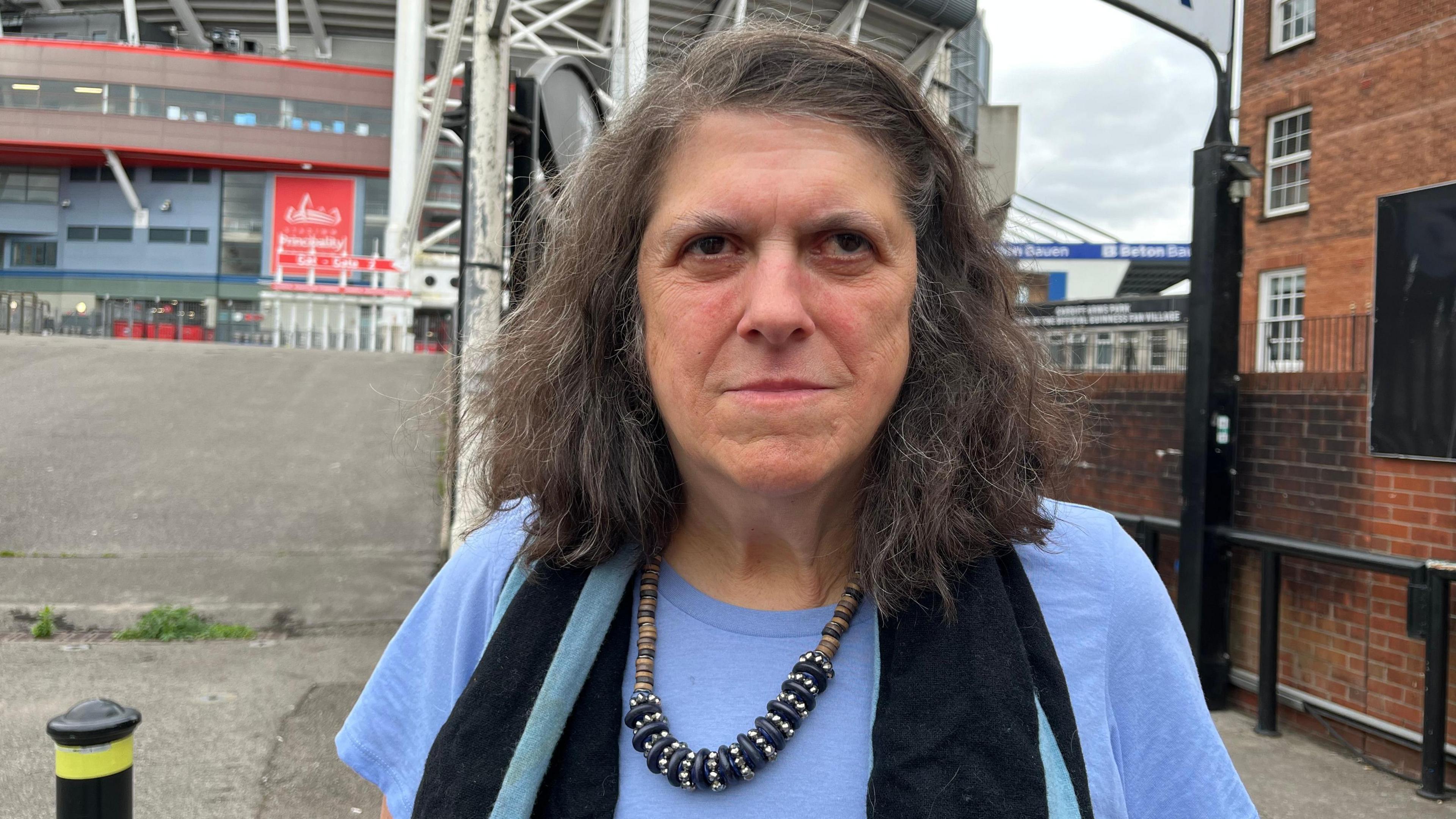 Close up of Lynn standing outside the Principality Stadium. She has medium length brown hair. Lynn is wearing a purple t-shirt, a black scarf and a beaded necklace.