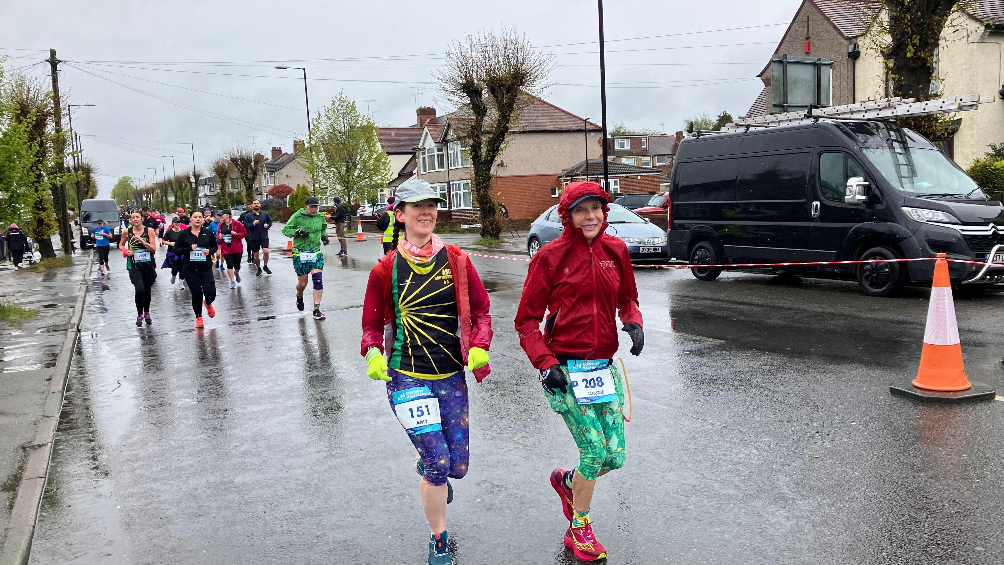 Two female runners wearing race numbers on their legs smile at the camera. The road surface they are running on is wet and more runners can be seen in the background, during an organised event. The woman on the let is wearing a cap and fluorescent yellow gloves, a black top with yellow and green pattern and star-patterned leggings. The women to her right is wearing a red jacket with hood up, green and pink leggings and red running shoes.