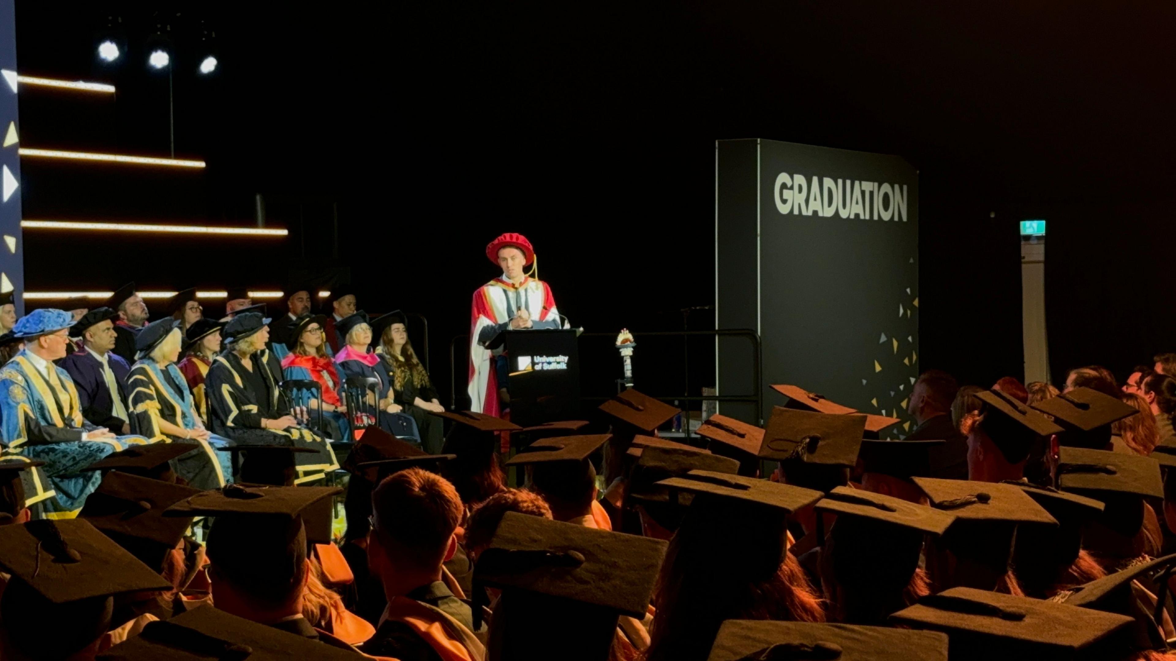 Kieran McKenna, wearing a red and yellow academic robe, stands at a lecturn in a hall. Sitting behind are a number of other people wearing academic robes. In front are the backs of heads wearing back mortar boards.