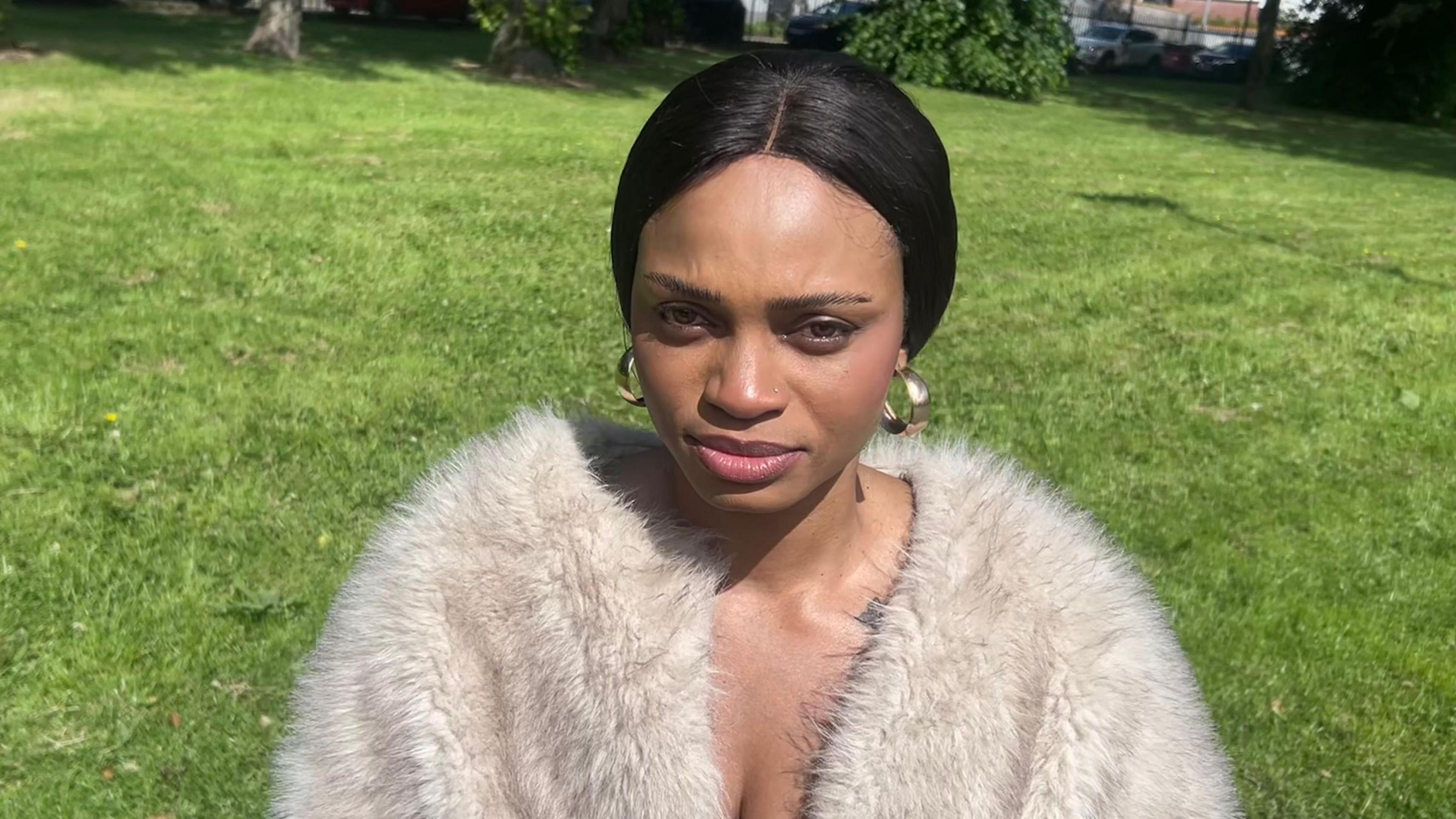 Chinonso looking at the camera. She has black hair and is wearing gold hoop earrings, a black top and cream fur jacket.